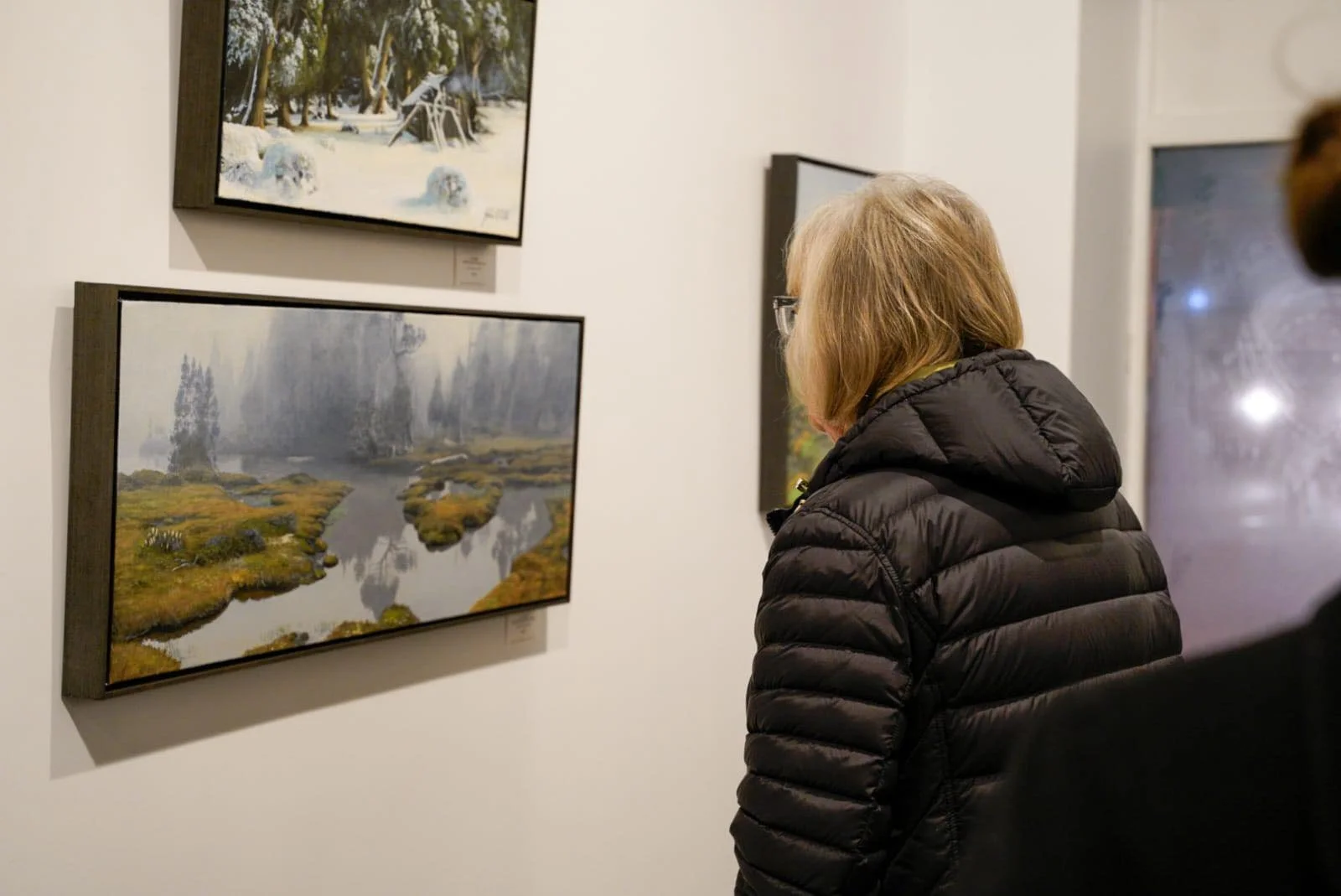 A woman with blonde hair and glasses, wearing a black puffy jacket, observes landscape paintings in an art gallery.