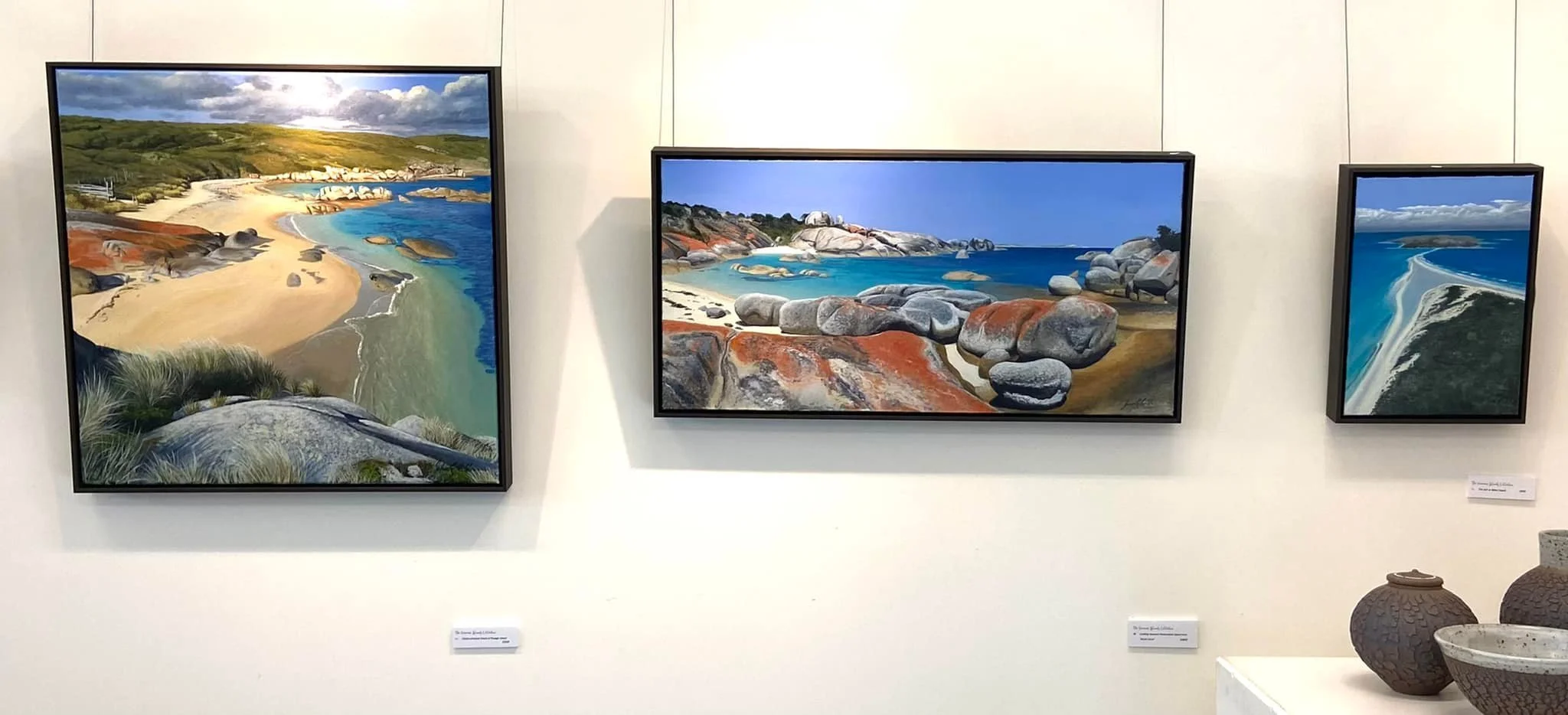 Three landscape paintings of coastal scenes displayed on a gallery wall, with pottery arranged below on a white table.