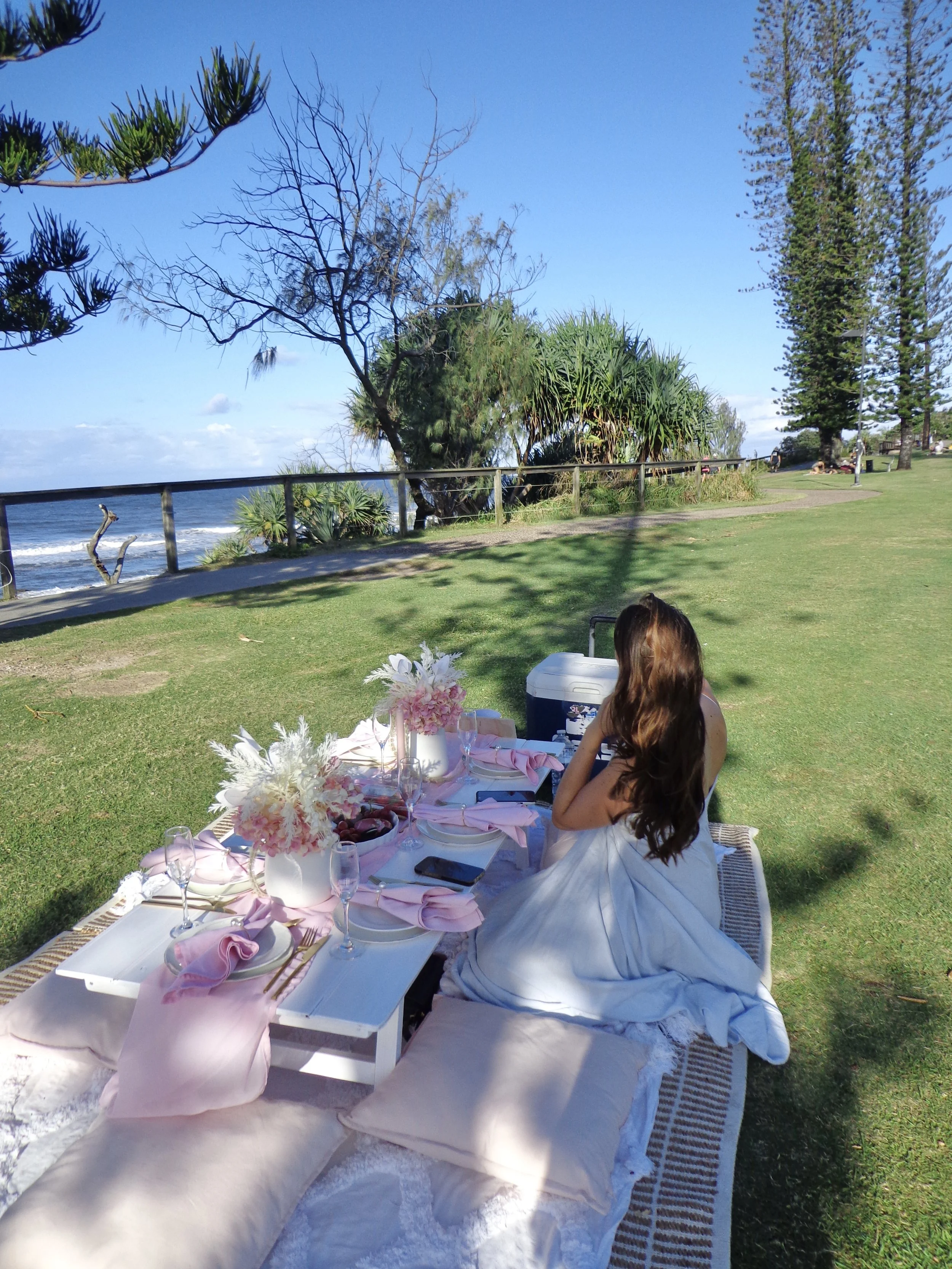 A woman sitting on a blanket during an outdoor picnic or gathering near the coast, with a table set with flowers, plates, and glasses, surrounded by grass, trees, and a view of the ocean.