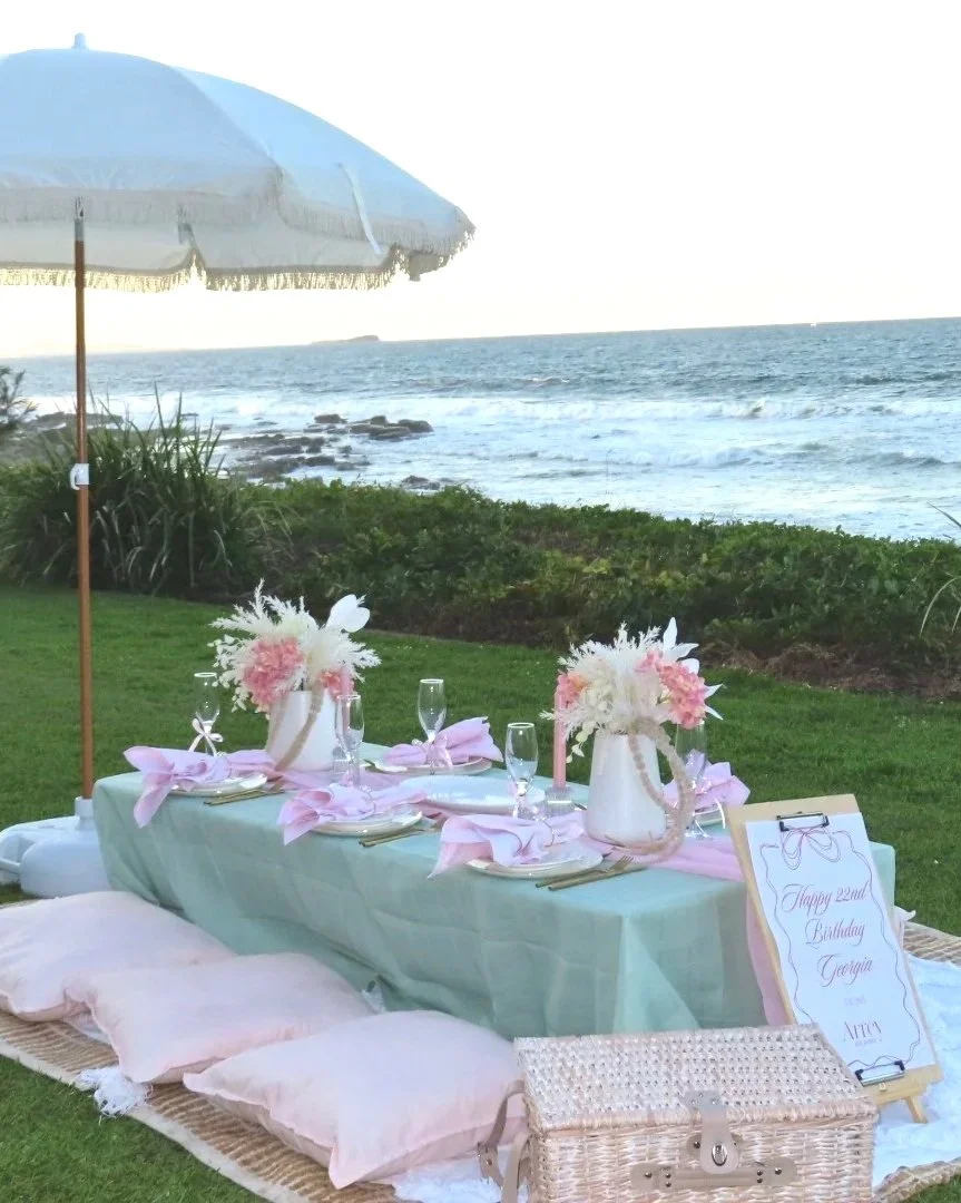 A seaside outdoor birthday celebration setup with a long table decorated with pink and white flowers in vases, light pink napkins, and champagne glasses. There are pink cushions on the ground and a large white umbrella providing shade. The table is set against a backdrop of the ocean and rocks.
