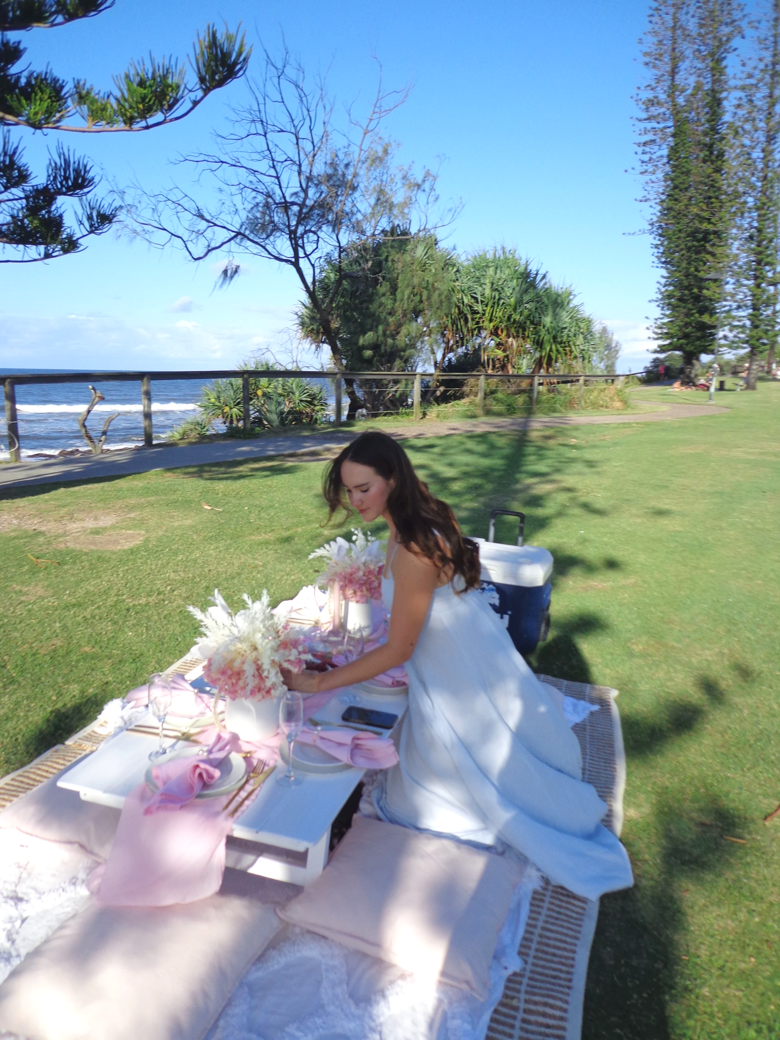 Young woman in a white dress setting up a picnic table outdoors by the ocean, with trees and a clear blue sky in the background.