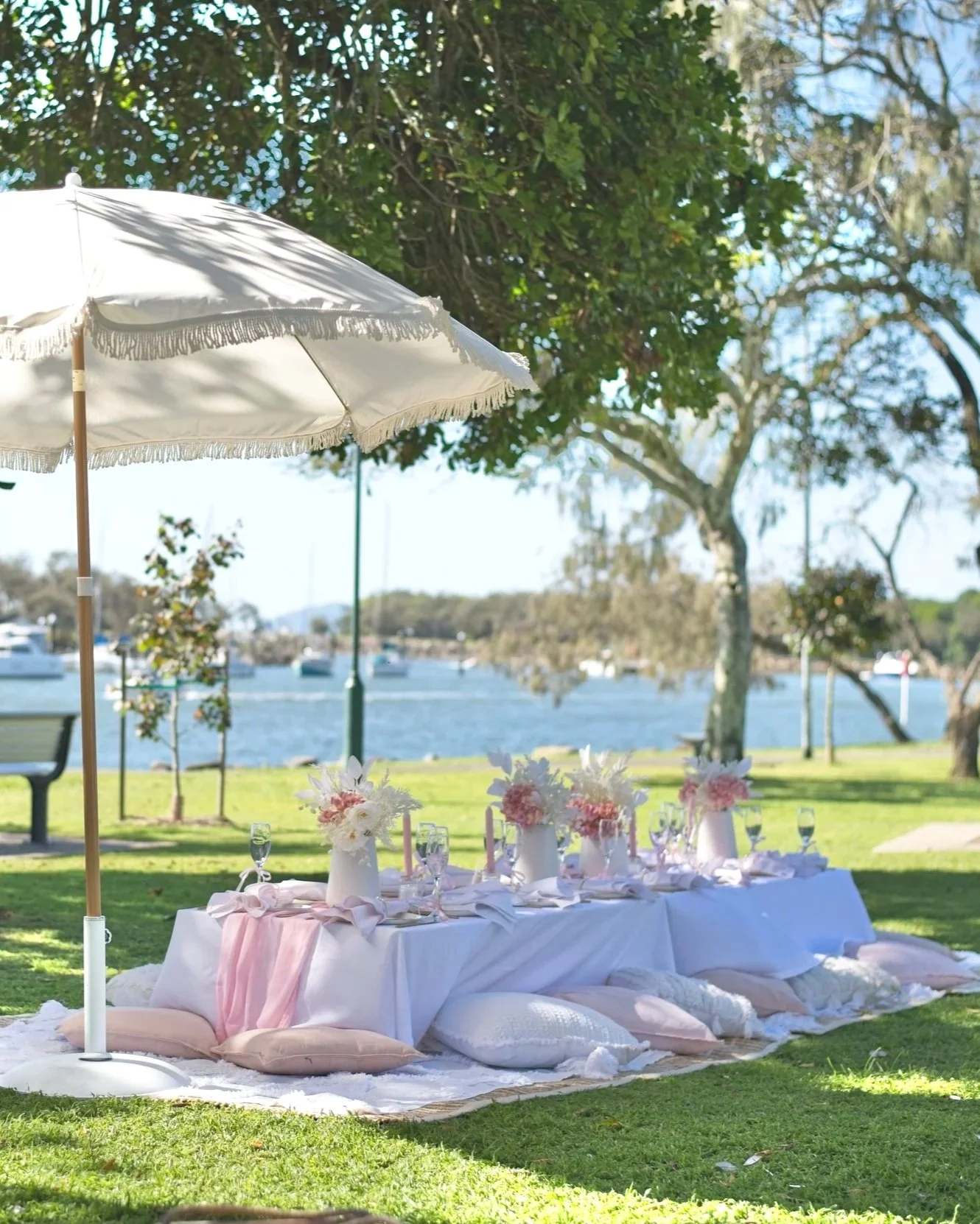 Decorated picnic table for outdoor birthday event near water with pink and white floral arrangements and cushions.