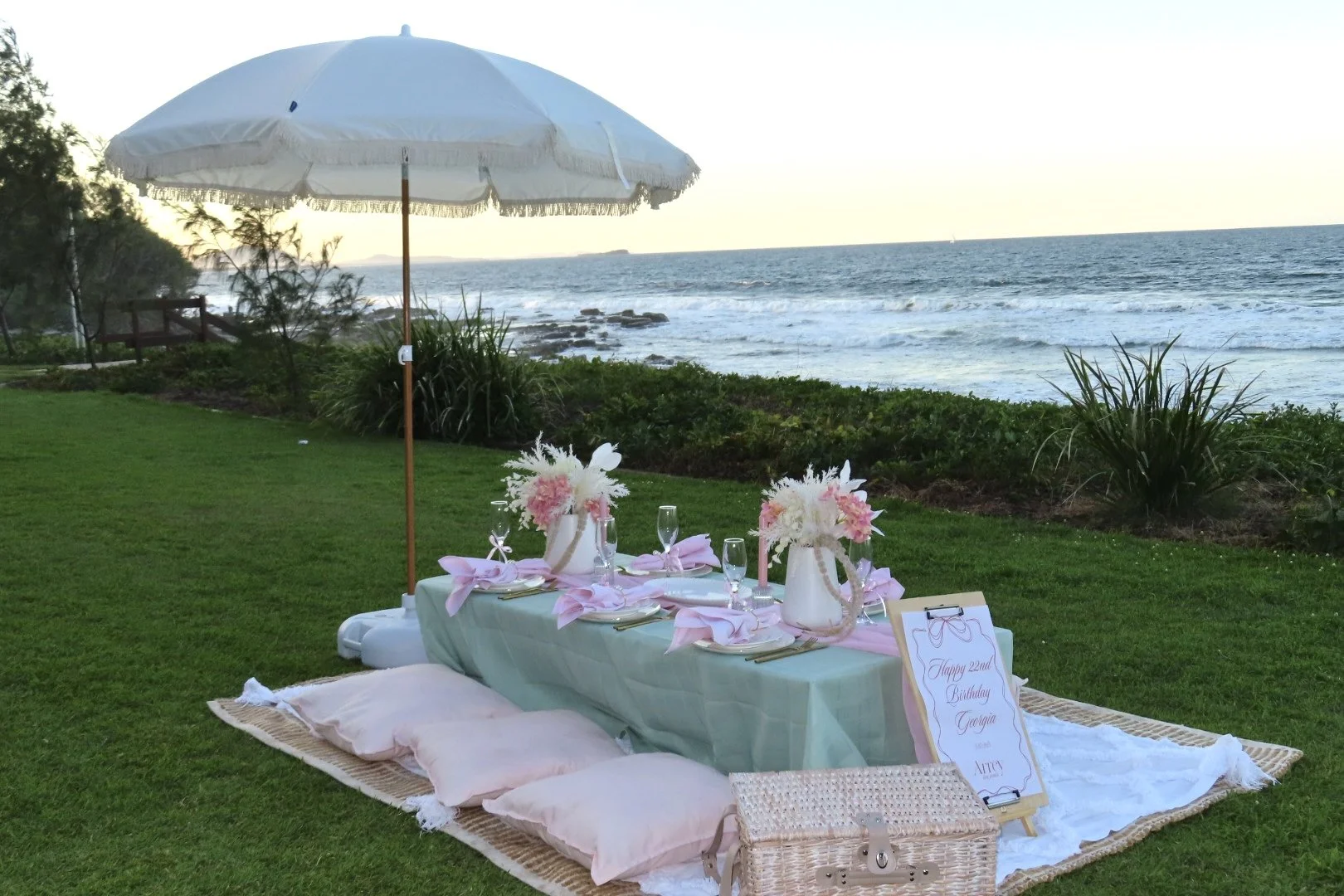 Beachside picnic setup with low table, pink cushions, floral arrangements, champagne glasses, and a large white umbrella overlooking the ocean at sunset.