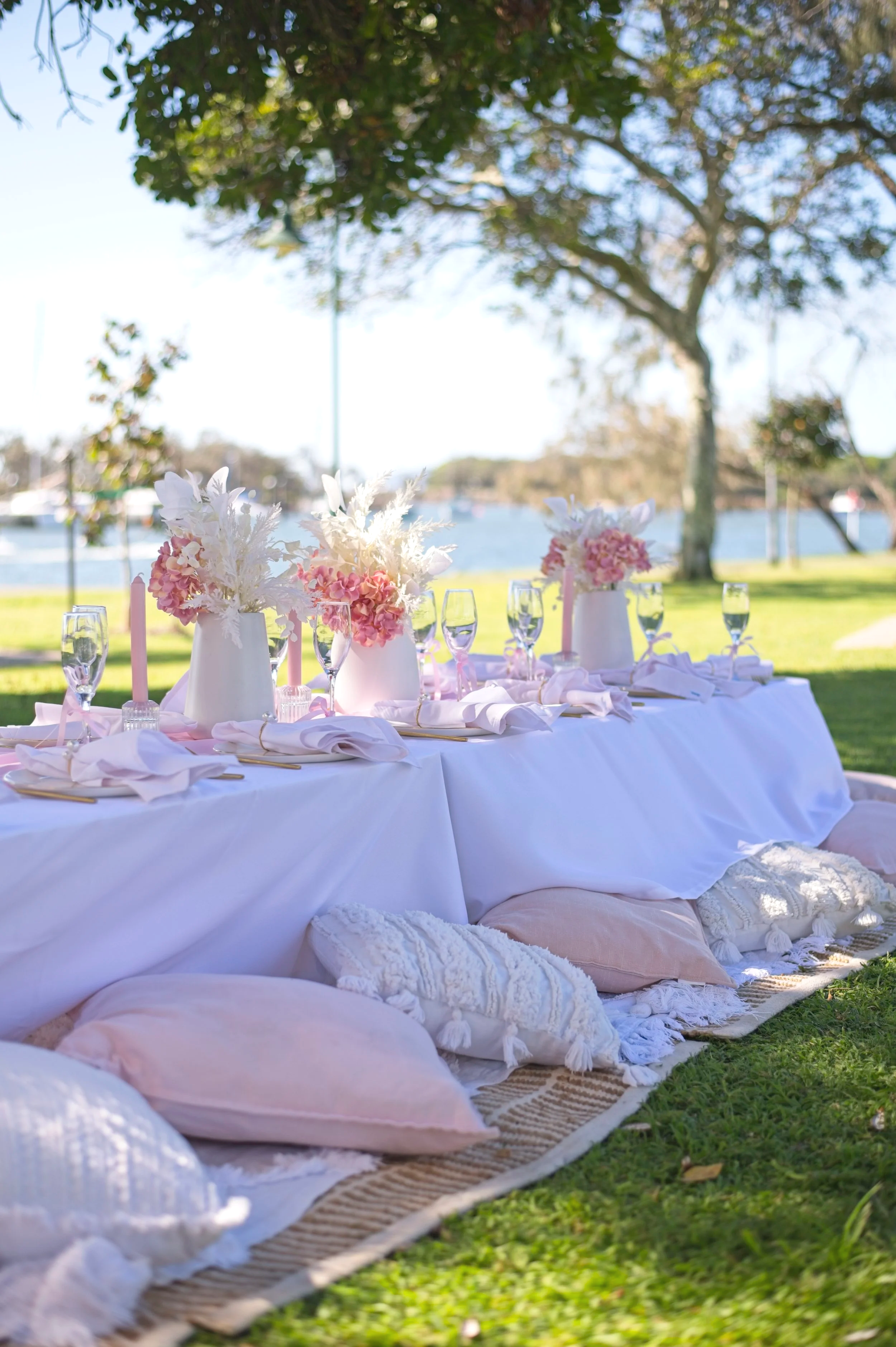 An outdoor dining setup featuring a long white tablecloth with elegant floral centerpieces, champagne glasses, and pink candles, set on a grassy area near a body of water with trees in the background.