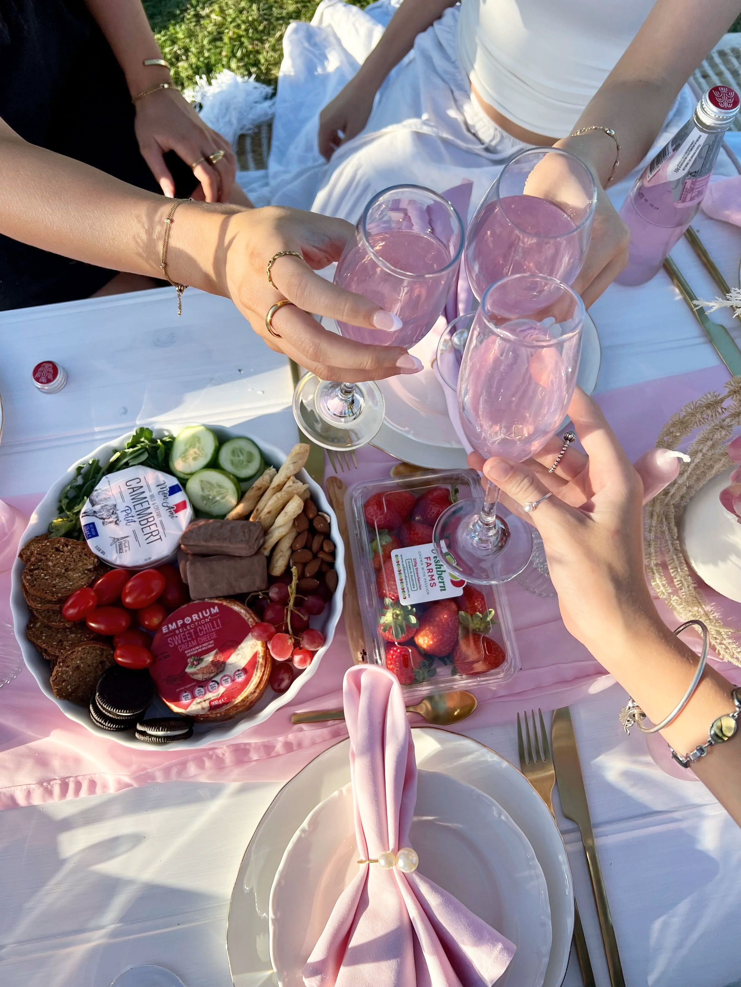 A group of people toasting with pink drinks at a table set for a celebration, with a platter of snacks and strawberries.