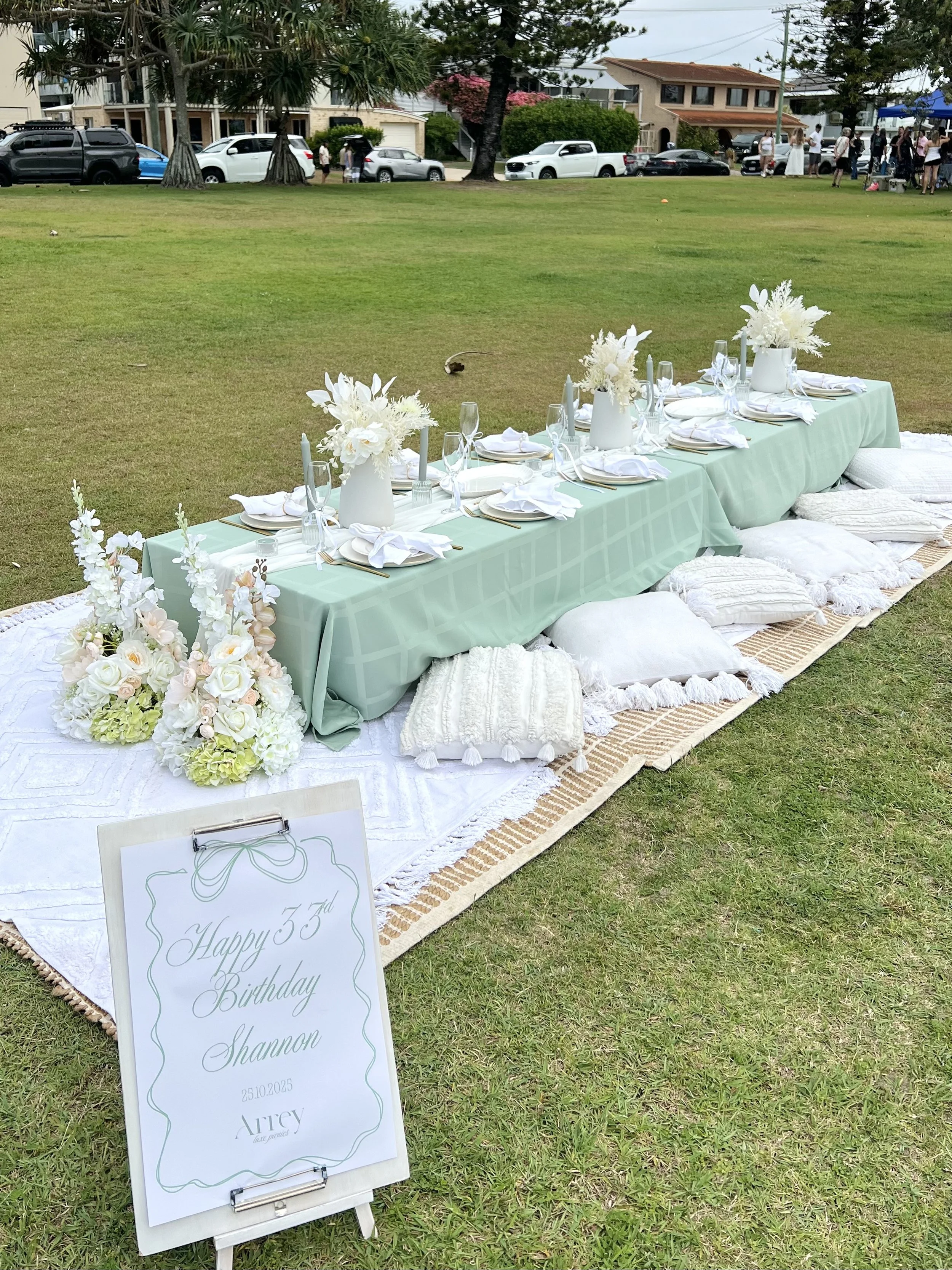 Outdoor birthday celebration with a long, green table set with white plates, napkins, glasses, and white floral centerpieces, placed on a white and beige patterned rug with cushions and pillows. A sign reads 'Happy 35th Birthday Shannon 25.10.2025'.