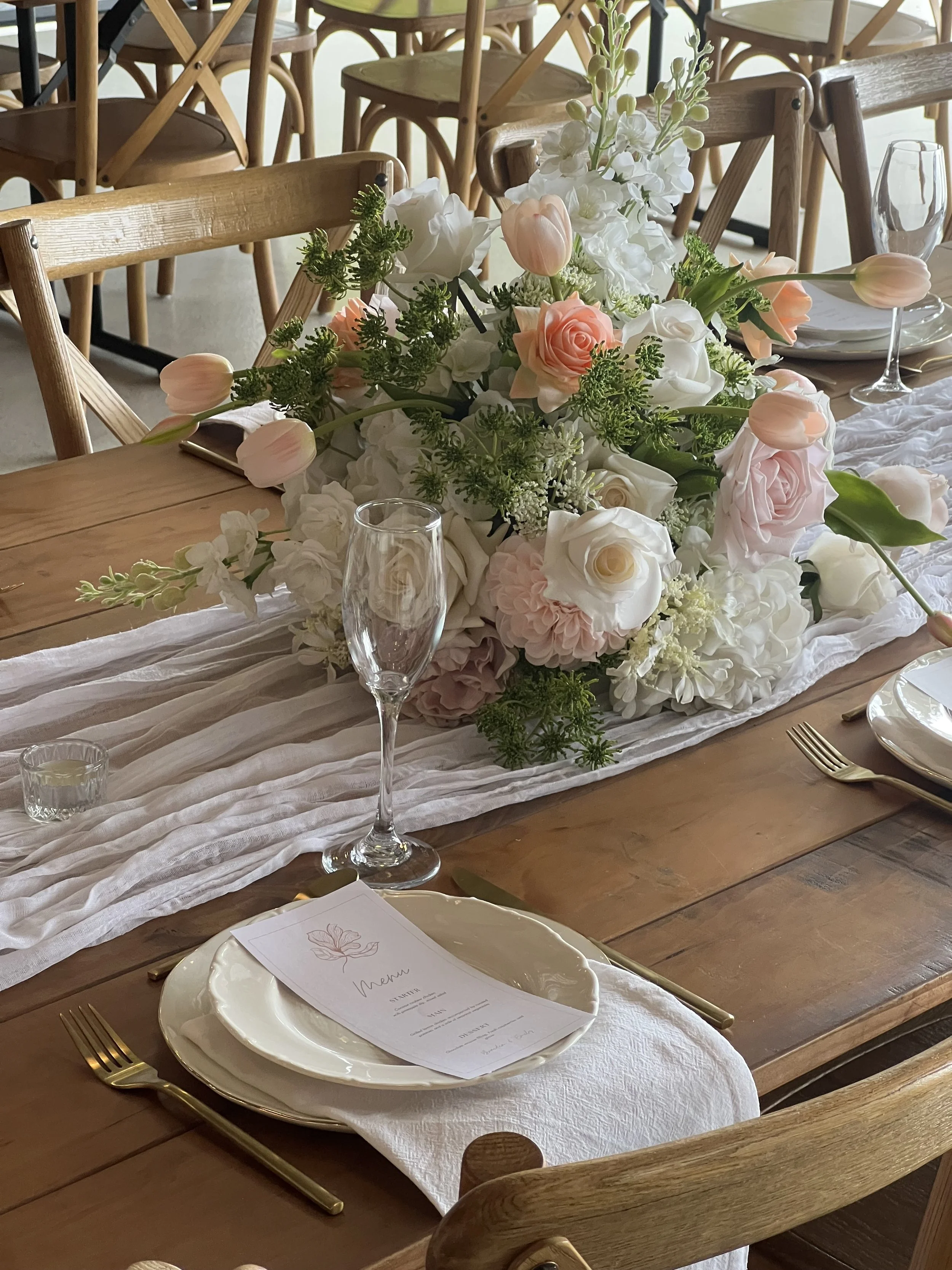 A table decorated with a large floral centerpiece of white and pink roses, tulips, and greenery. The table has a white cloth runners, plates, gold utensils, and a menu, with empty glasses set for a formal event.