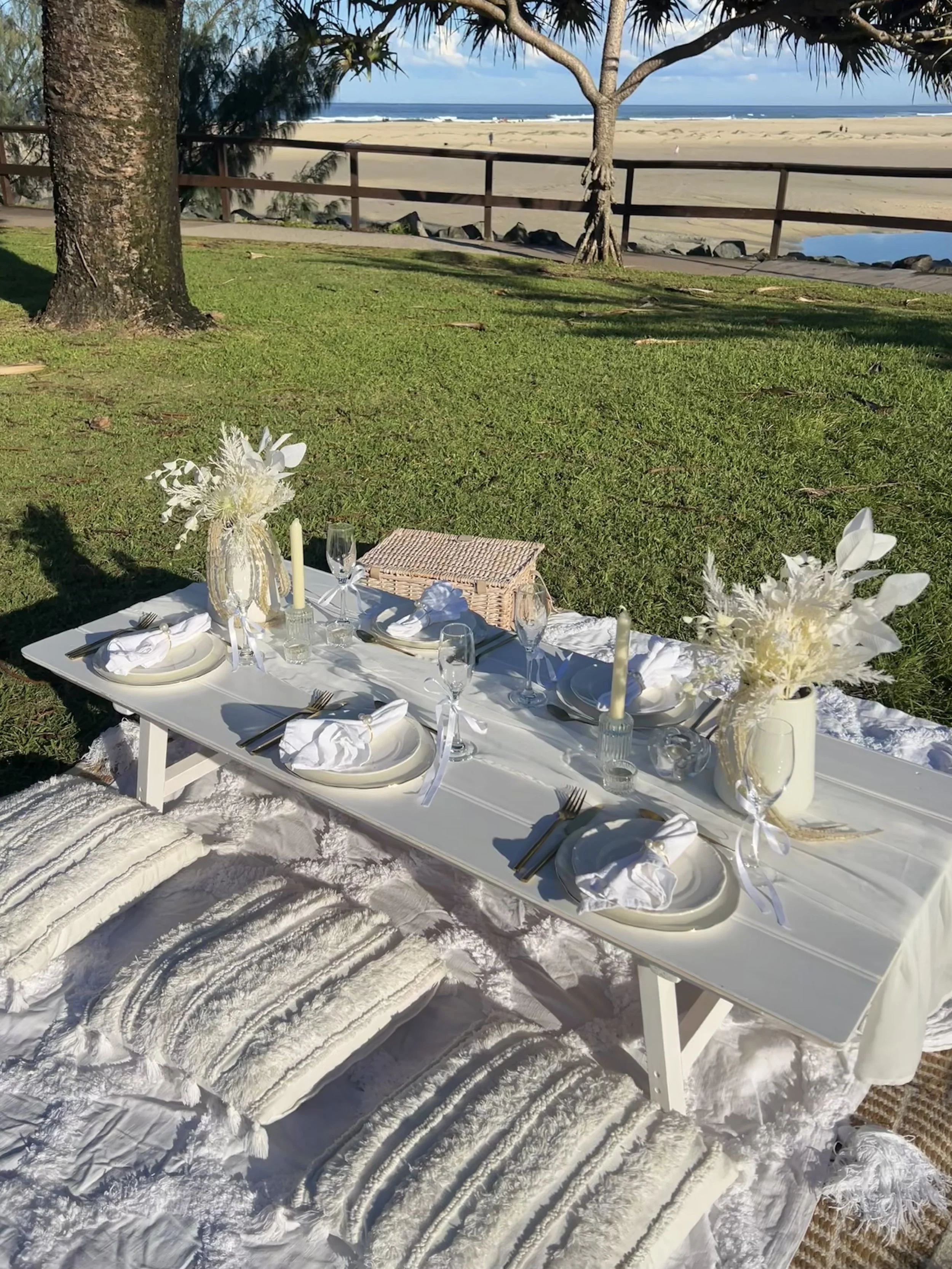 A picnic-style dining setup on the grass near a beach, with a white table, white floral arrangements, candles, wine glasses, and place settings, with a view of the ocean and beach in the background.