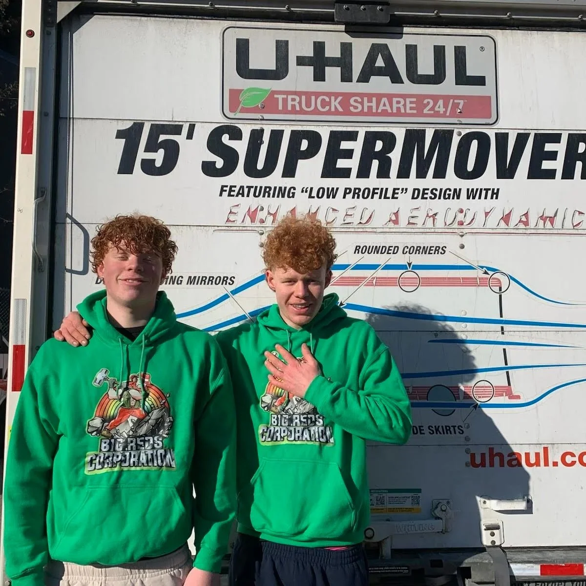 Two young men with curly red hair wearing matching green hoodies with a graphic design for Big Reds Corporation, standing in front of a U-Haul truck with promotional signage for a 15-foot super mover