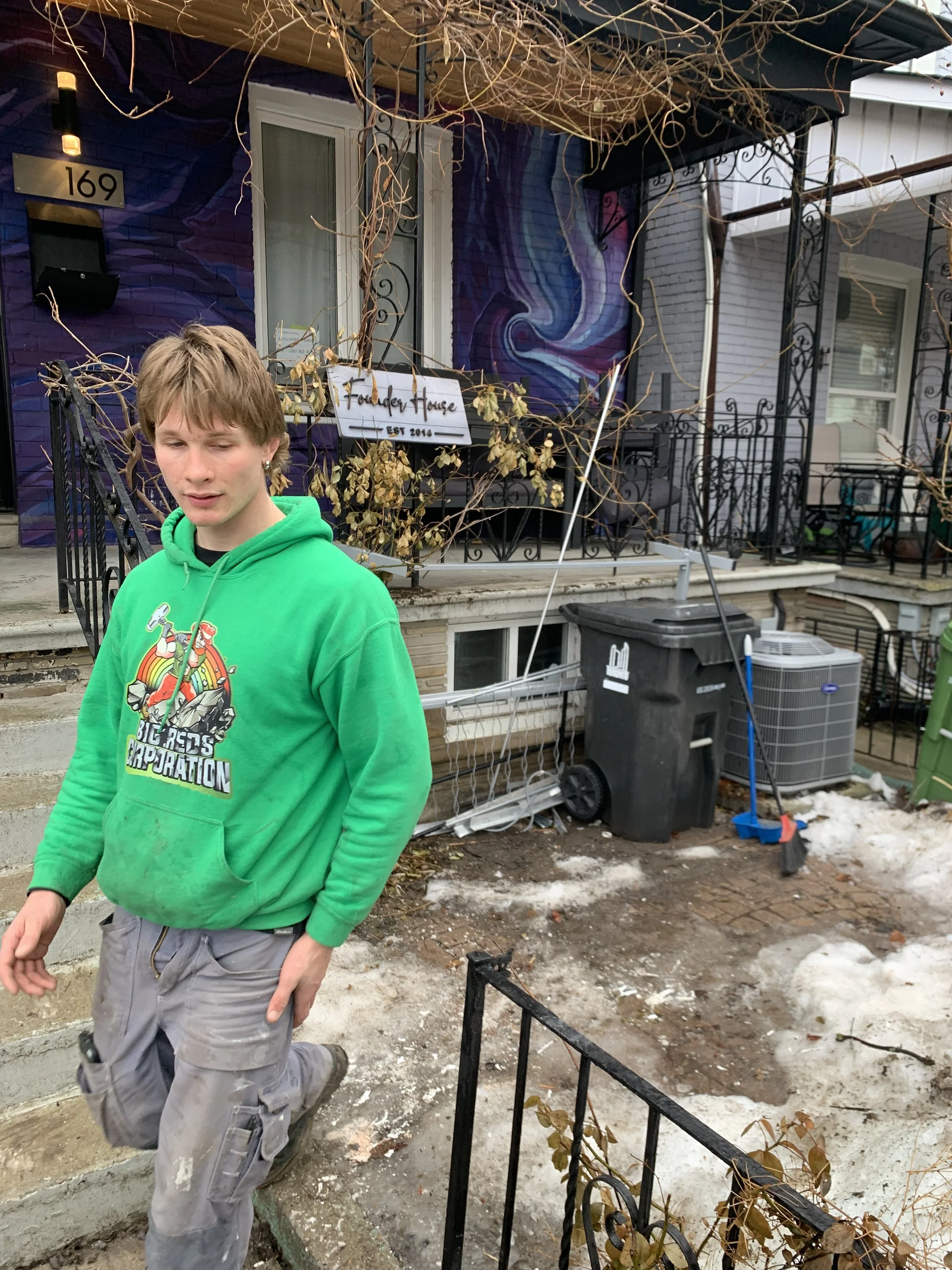 A young man in a bright green hoodie and gray shorts is walking outside. Behind him, there is a house with a purple and blue mural, leafless vines, and outdoor furniture. There is snow on the ground.