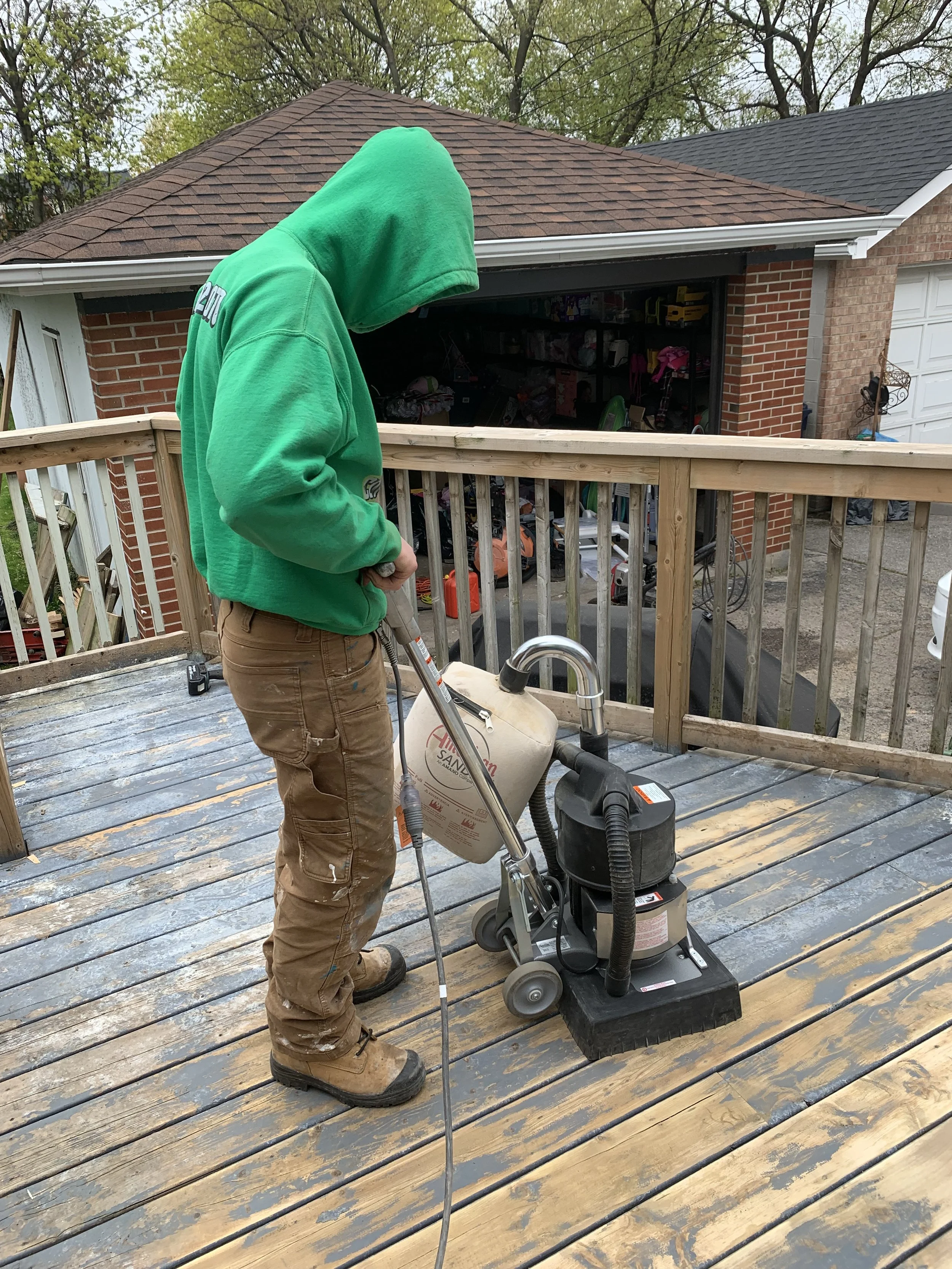 A man in a green hoodie and brown cargo pants operating a wet/dry vacuum on a wooden deck outside a house with a garage and brick walls.