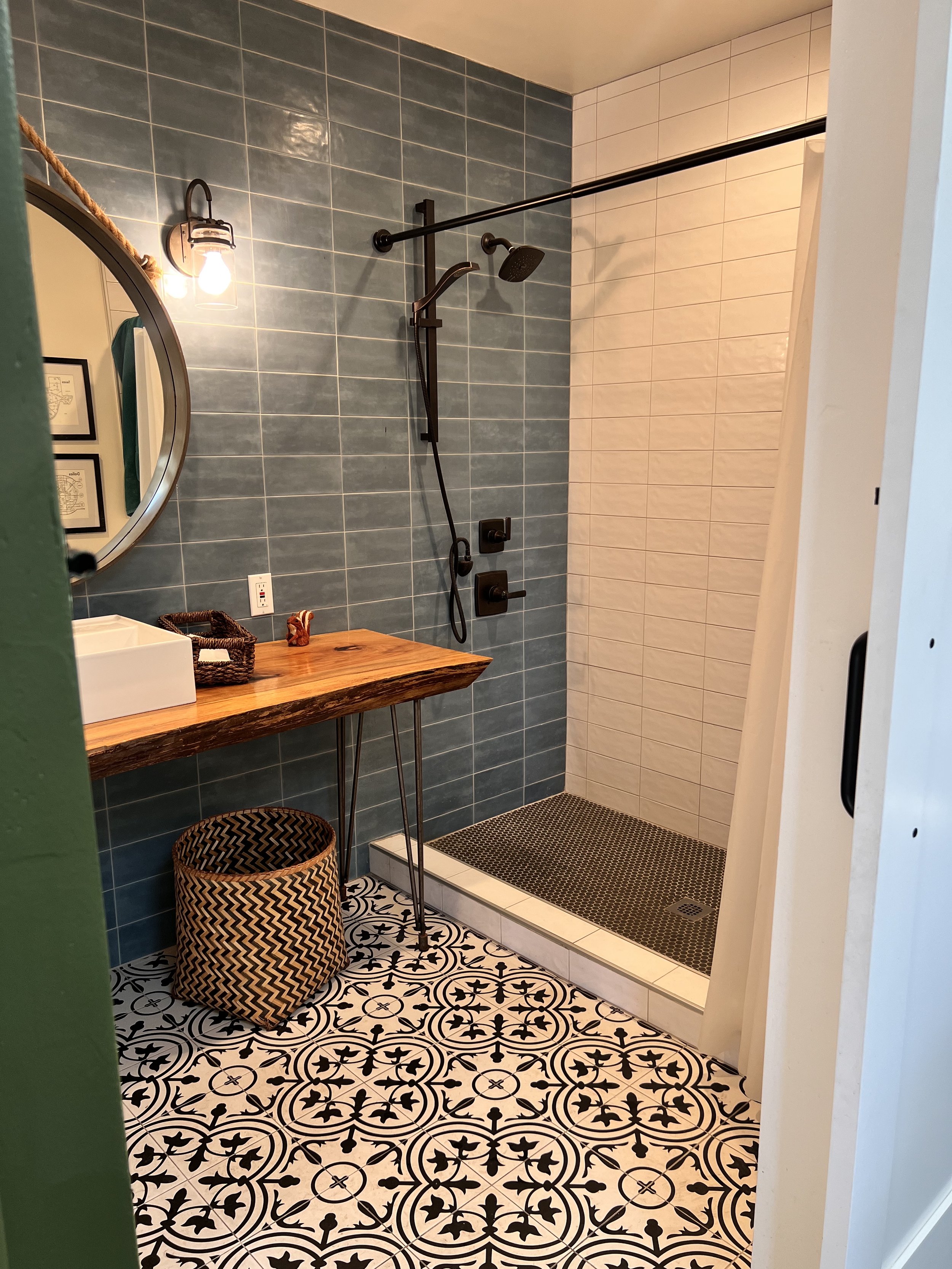A modern bathroom with a walk-in shower, featuring blue tiles on one wall and white tiles on the others. The shower has black fixtures and a black showerhead. There is a round mirror above a rustic wooden vanity with a sink, a woven basket on the flo