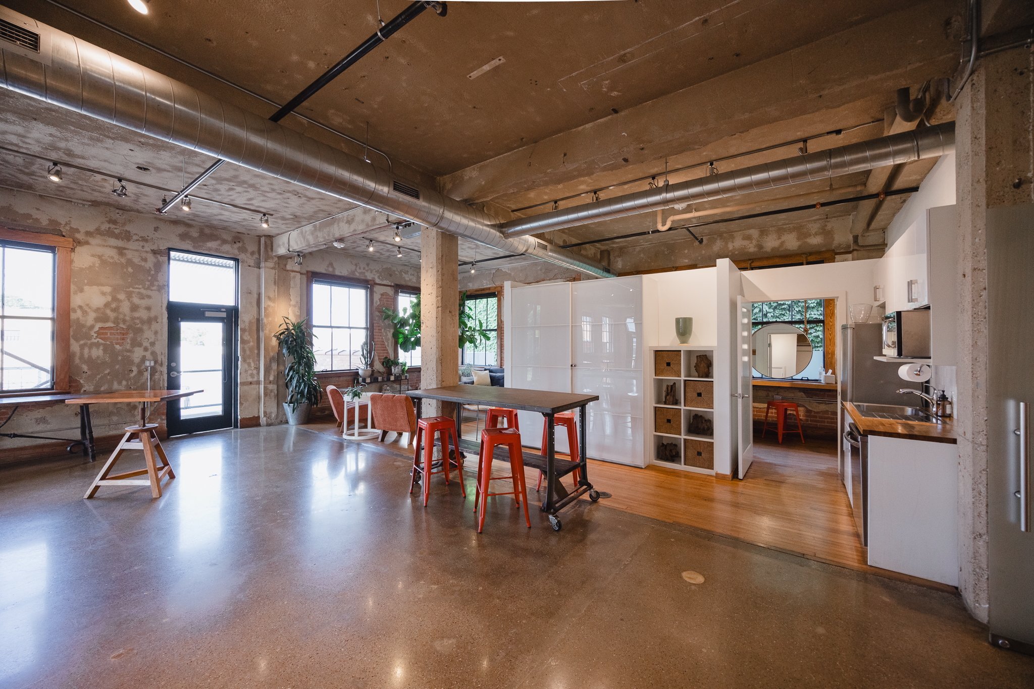 Inside a modern loft-style apartment with exposed ductwork and brick walls, featuring large windows, a wooden floor area with white walls, a kitchenette, and various furniture including chairs, tables, and plants.