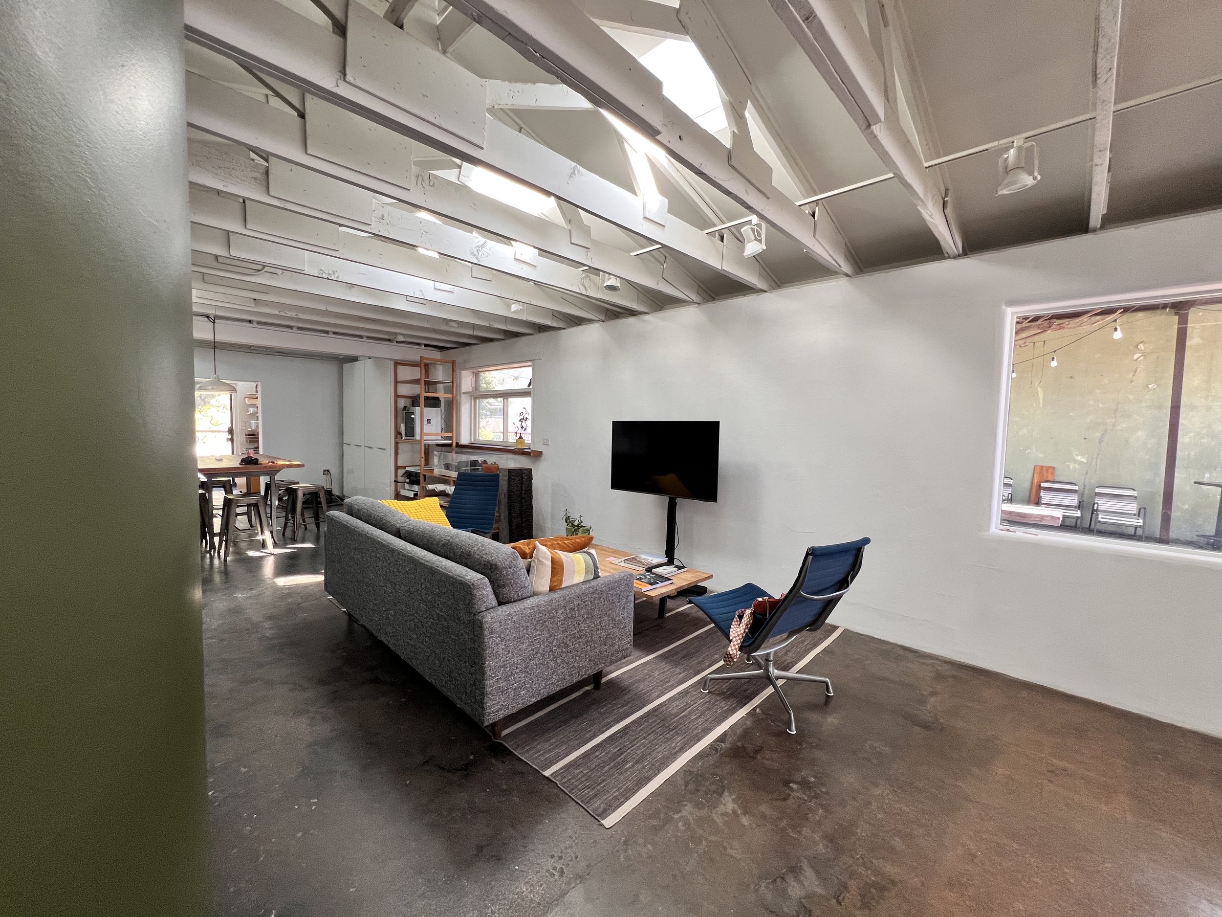 Open-concept living room with gray sofa, two blue chairs, a flat-screen TV on the wall, wooden coffee table, and dining area in the background with a wooden table and chairs, in a modern industrial style space with white walls and exposed ceiling bea