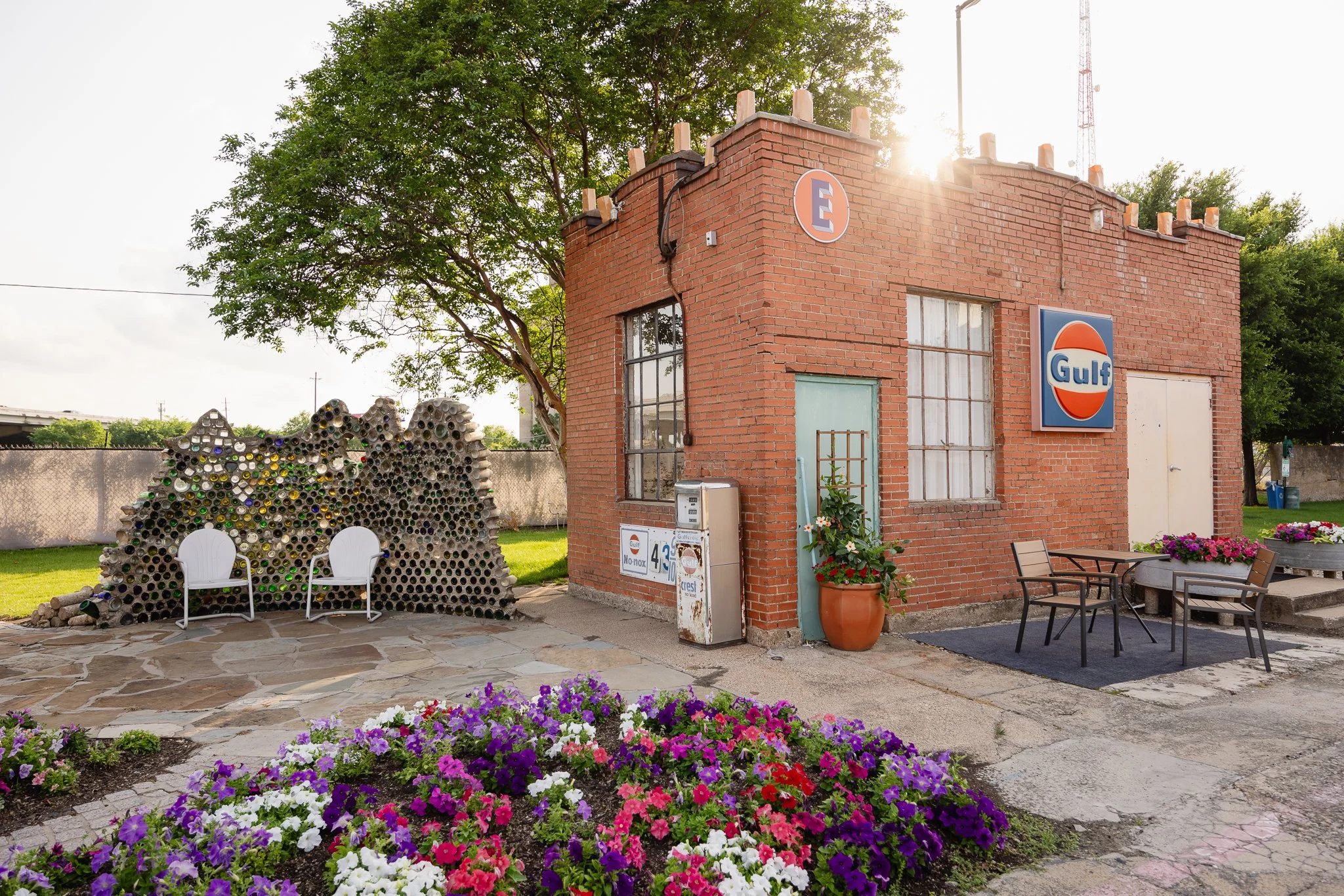 A small brick building with a Gulf logo sign on the front, surrounded by flowerbeds with colorful flowers, outdoor chairs, and a unique sculpture made of rolled-up bottles on the left side. The building has a green door, large windows, and a parking 