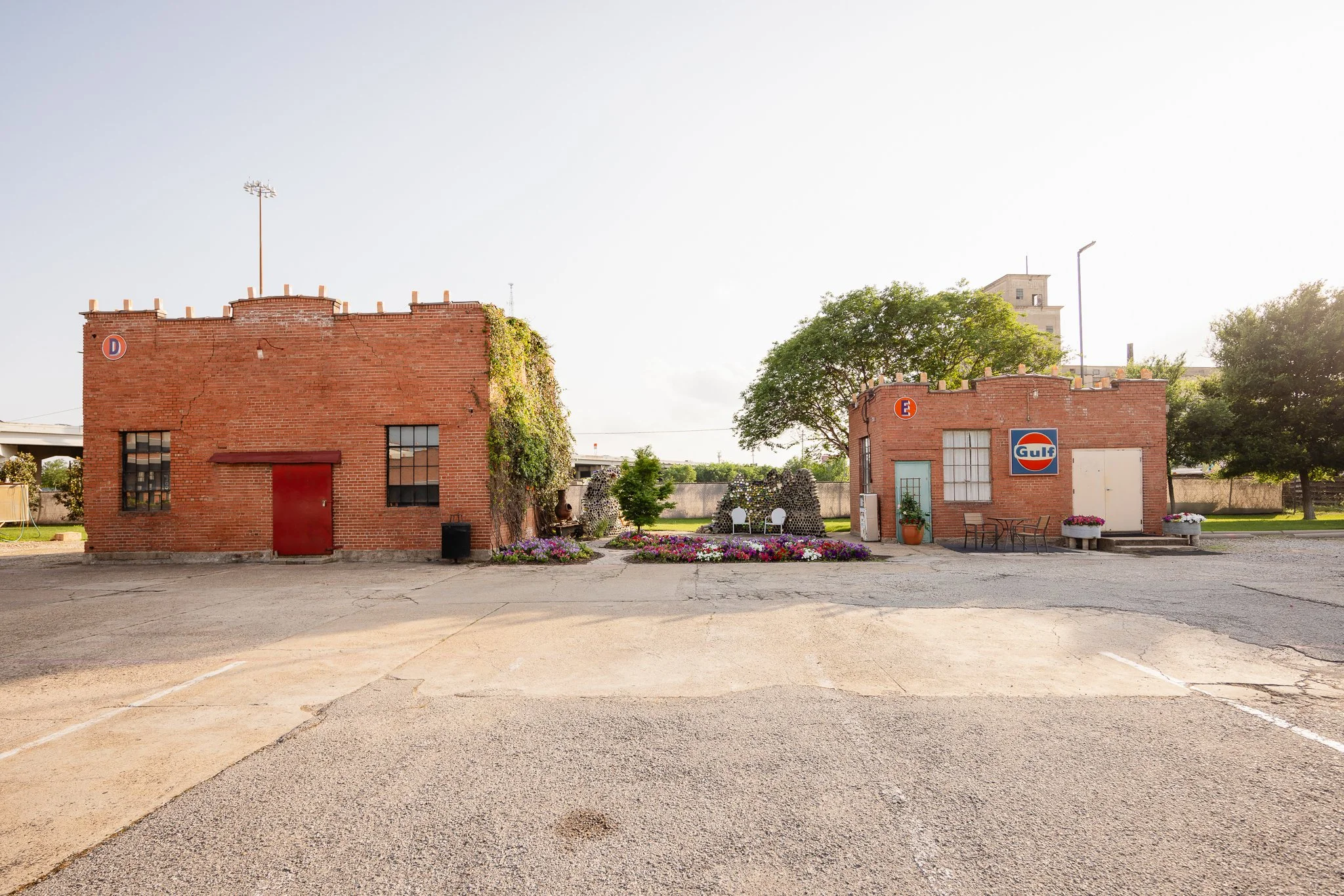 Two small red-brick buildings with vintage signage, potted plants, and outdoor seating, set against a park with trees and a clear sky.