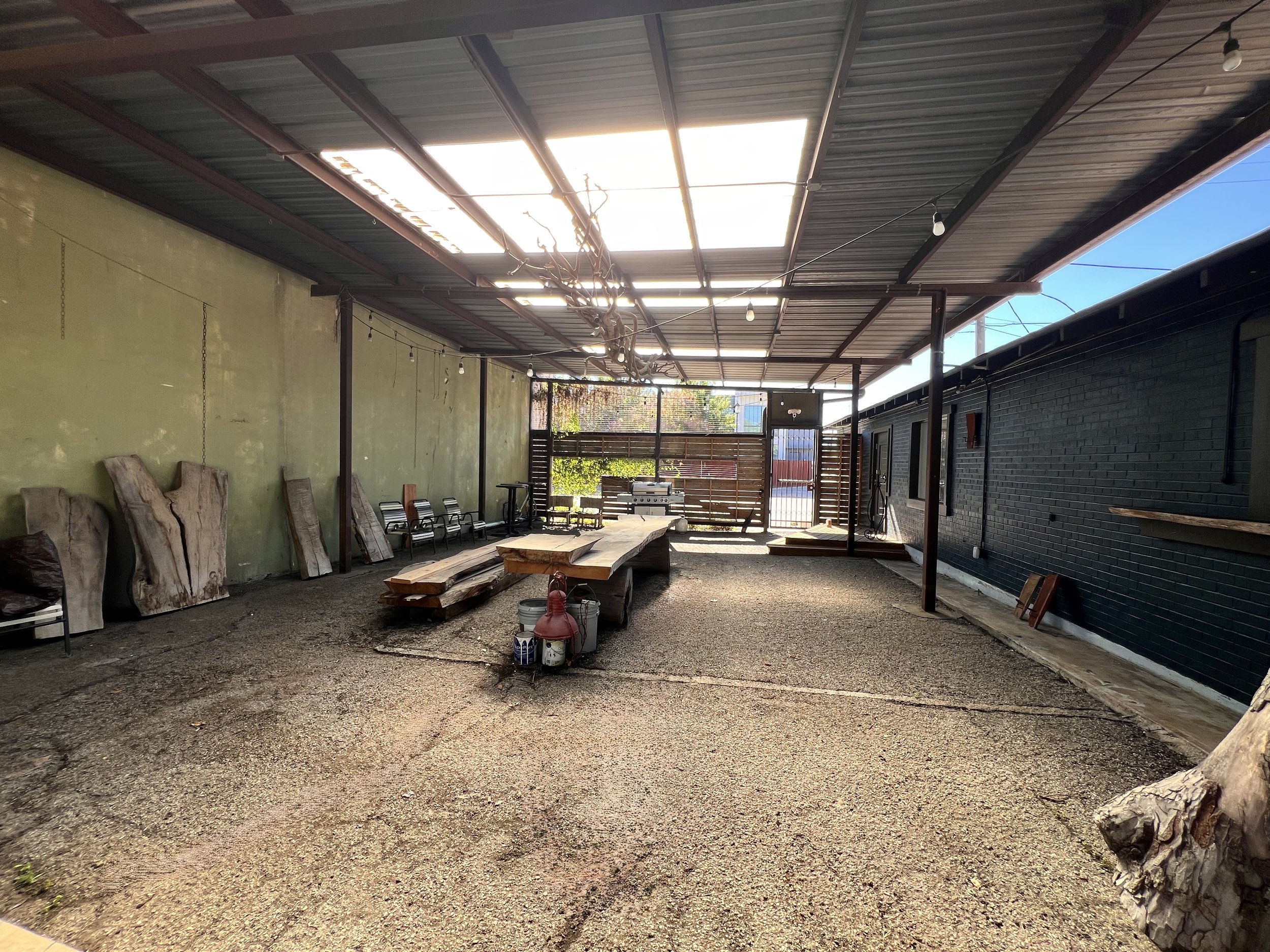 An outdoor patio area with a covered roof, a long wooden table with benches, metal chairs, cut wooden slabs, and a grill at the back. There is gravel on the ground and a partially painted green wall on the left side.