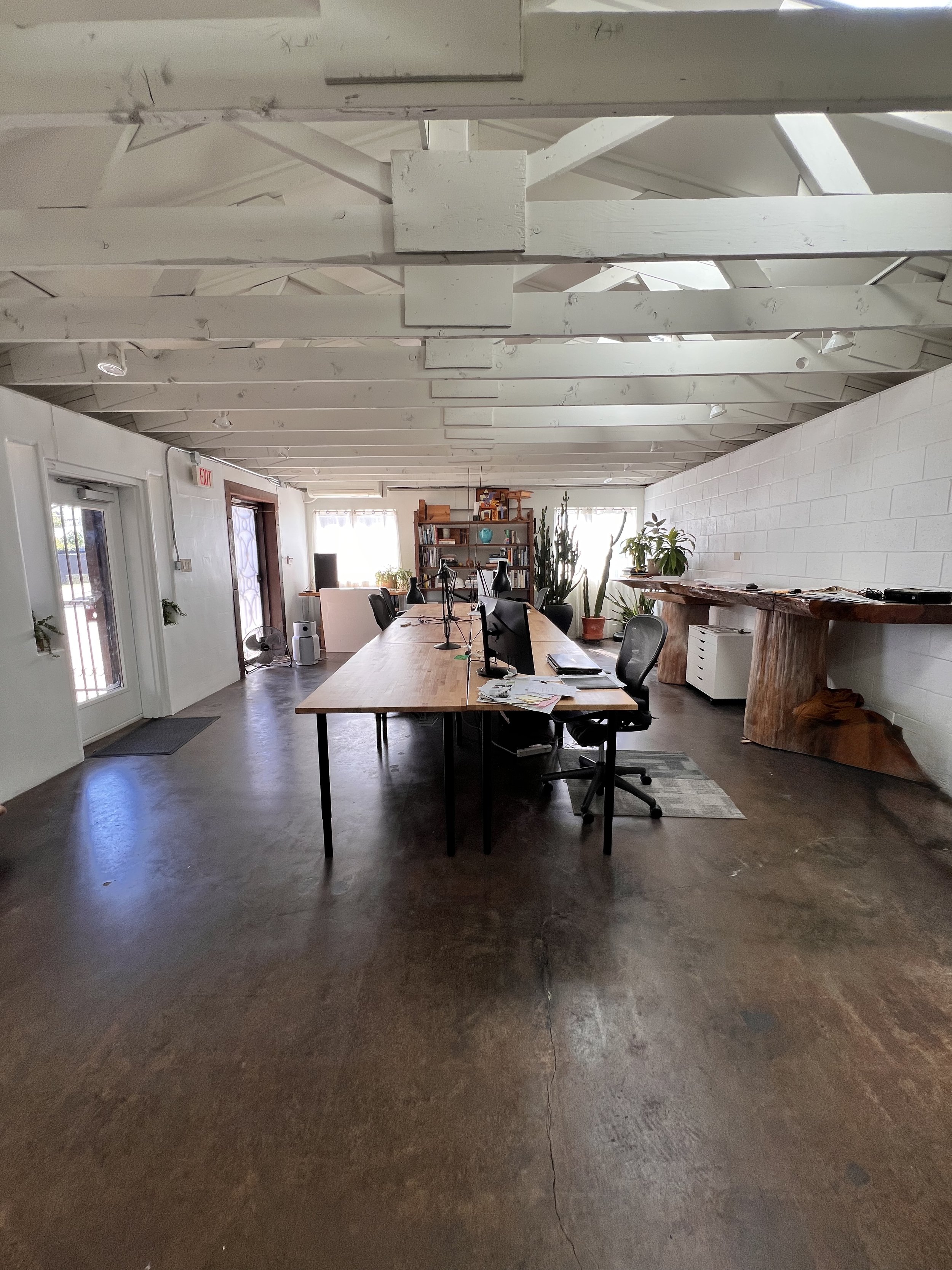 An office with a long wooden table, multiple chairs, and various office supplies. There are large windows with blinds, plants along the wall, and a light-colored brick wall with a rustic wooden shelf. The ceiling is exposed with white beams, and the 