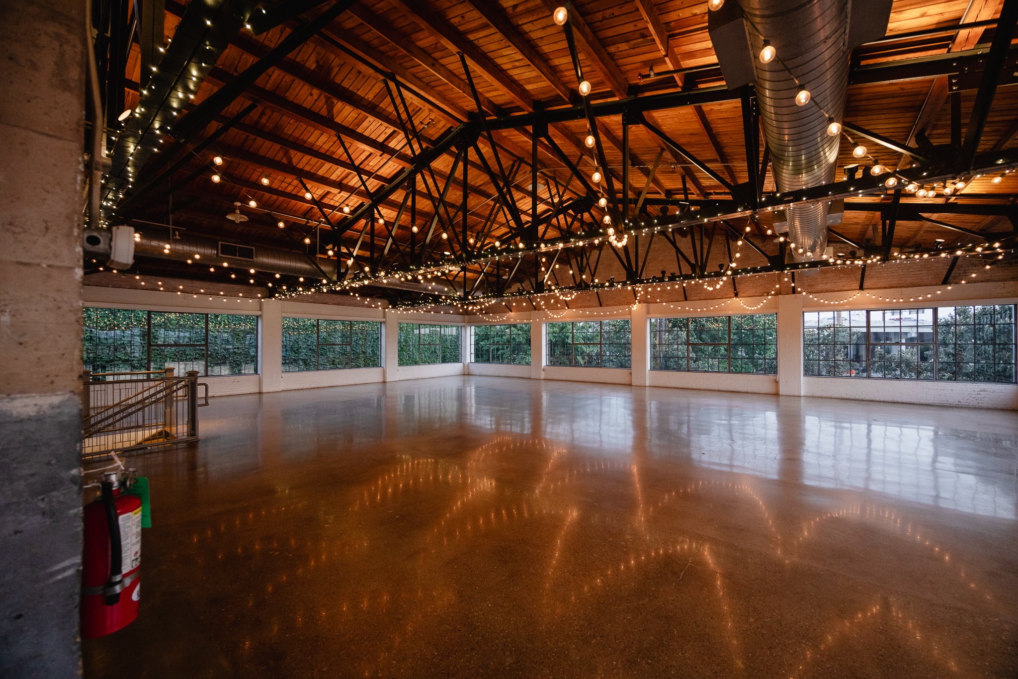 Empty event space with polished wooden floor, string lights, and large windows with greenery outside.