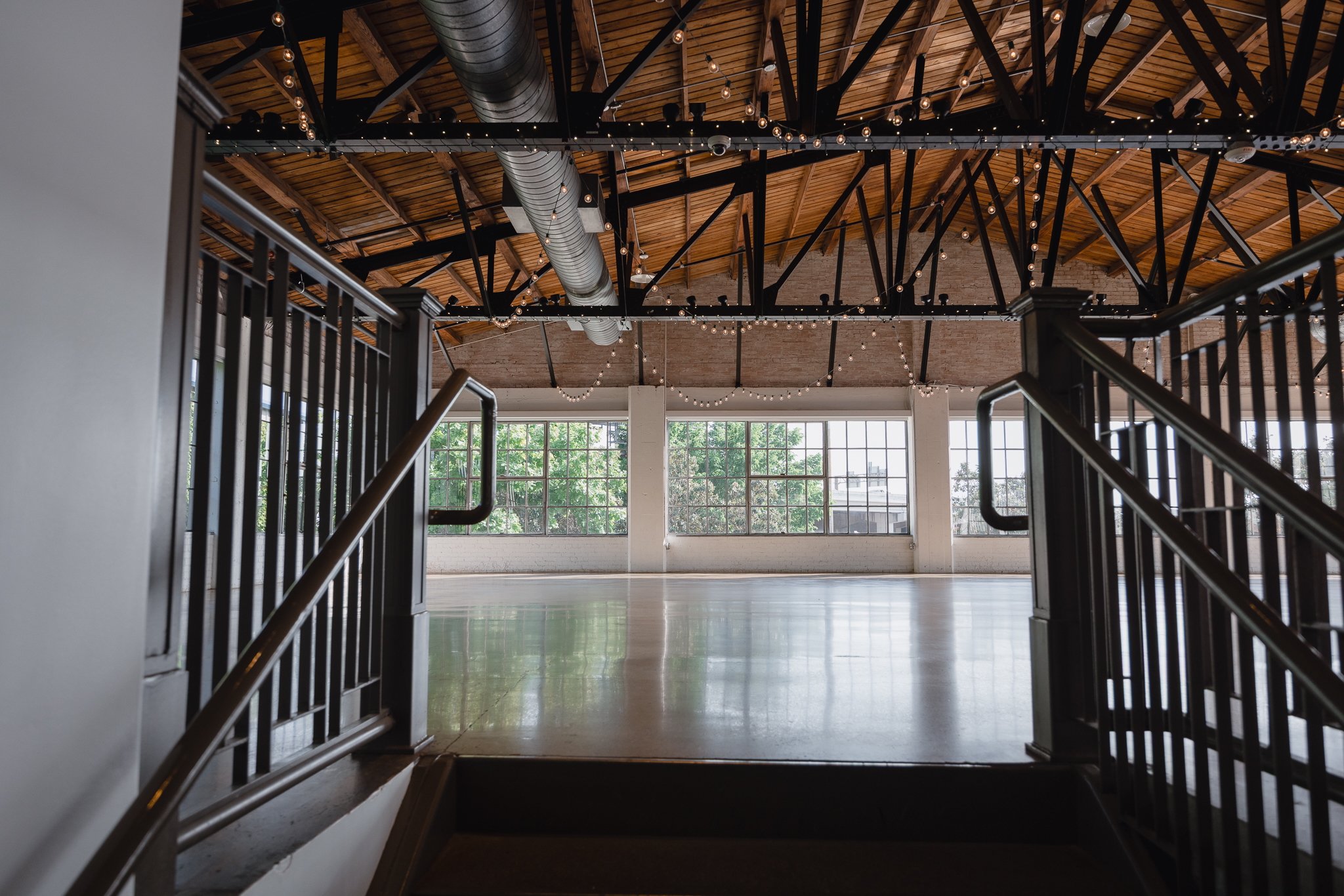 Empty indoor event space with large windows, exposed brick walls, a polished floor, string lights hanging from the wooden ceiling, and black metal railings.