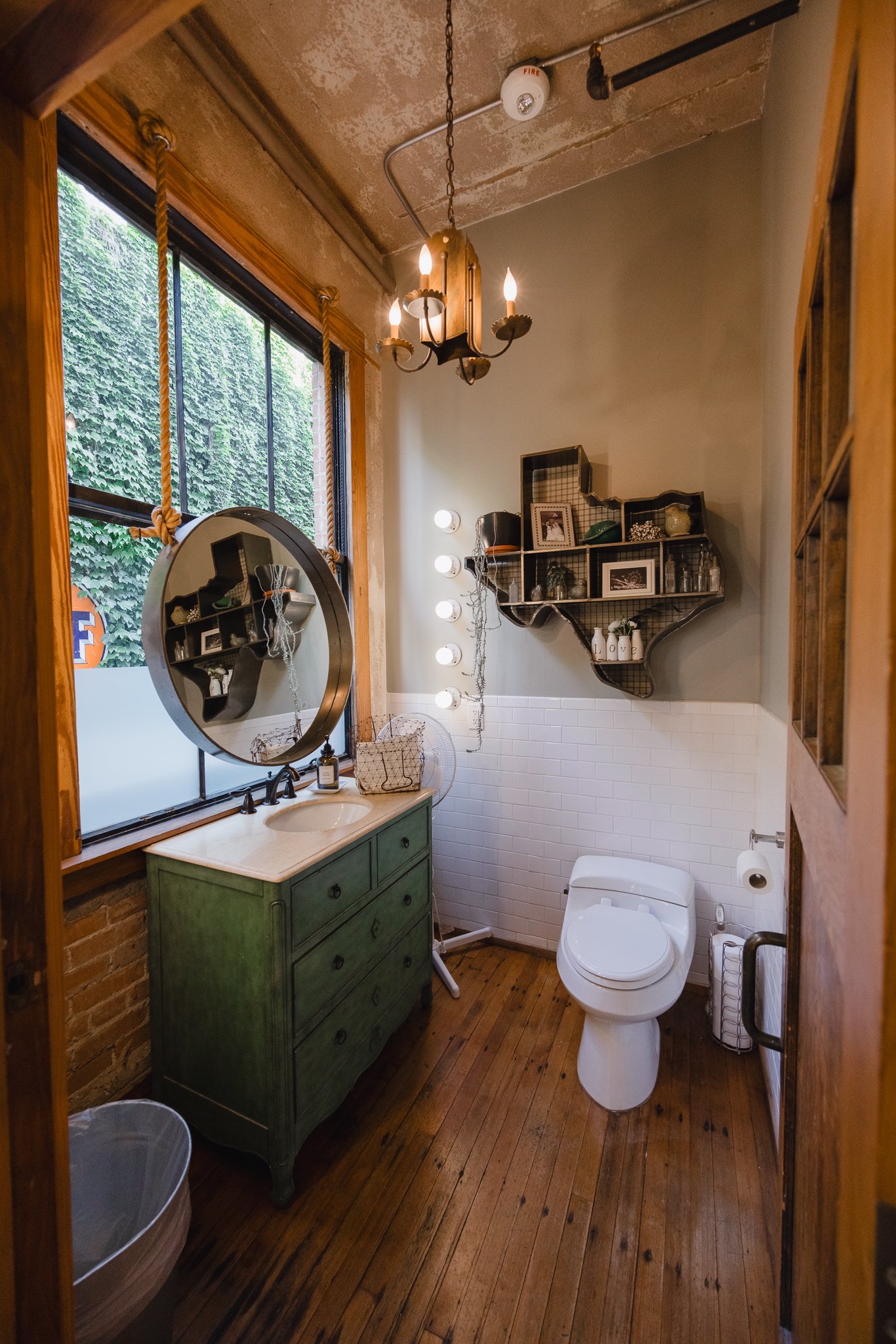 A small bathroom with wooden floors, a green dresser with a marble top, a round mirror, a window with wooden trim, a modern toilet, a wire wall shelf with decorative items, and a chandelier with candles hanging from the ceiling.