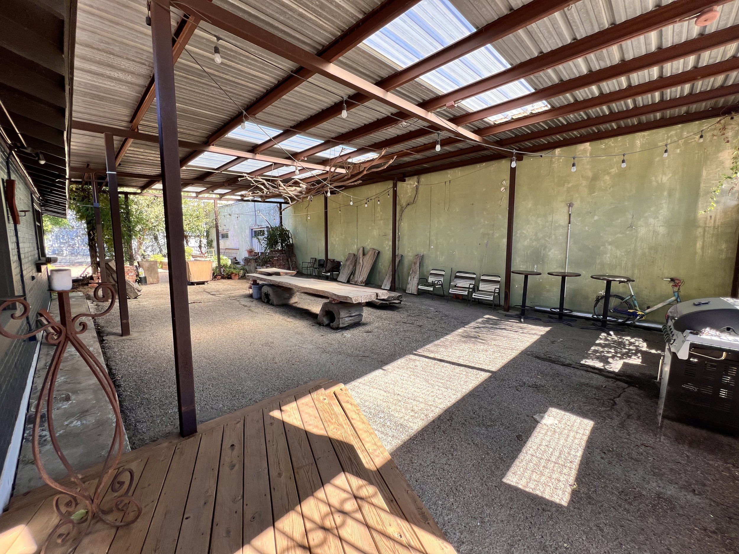 Outdoor covered patio area with gravel ground, wooden deck, and string lights hanging from metal beams. There are chairs, tables, potted plants, a bicycle, and a grill in the space. Sunlight creates shadows on the ground.