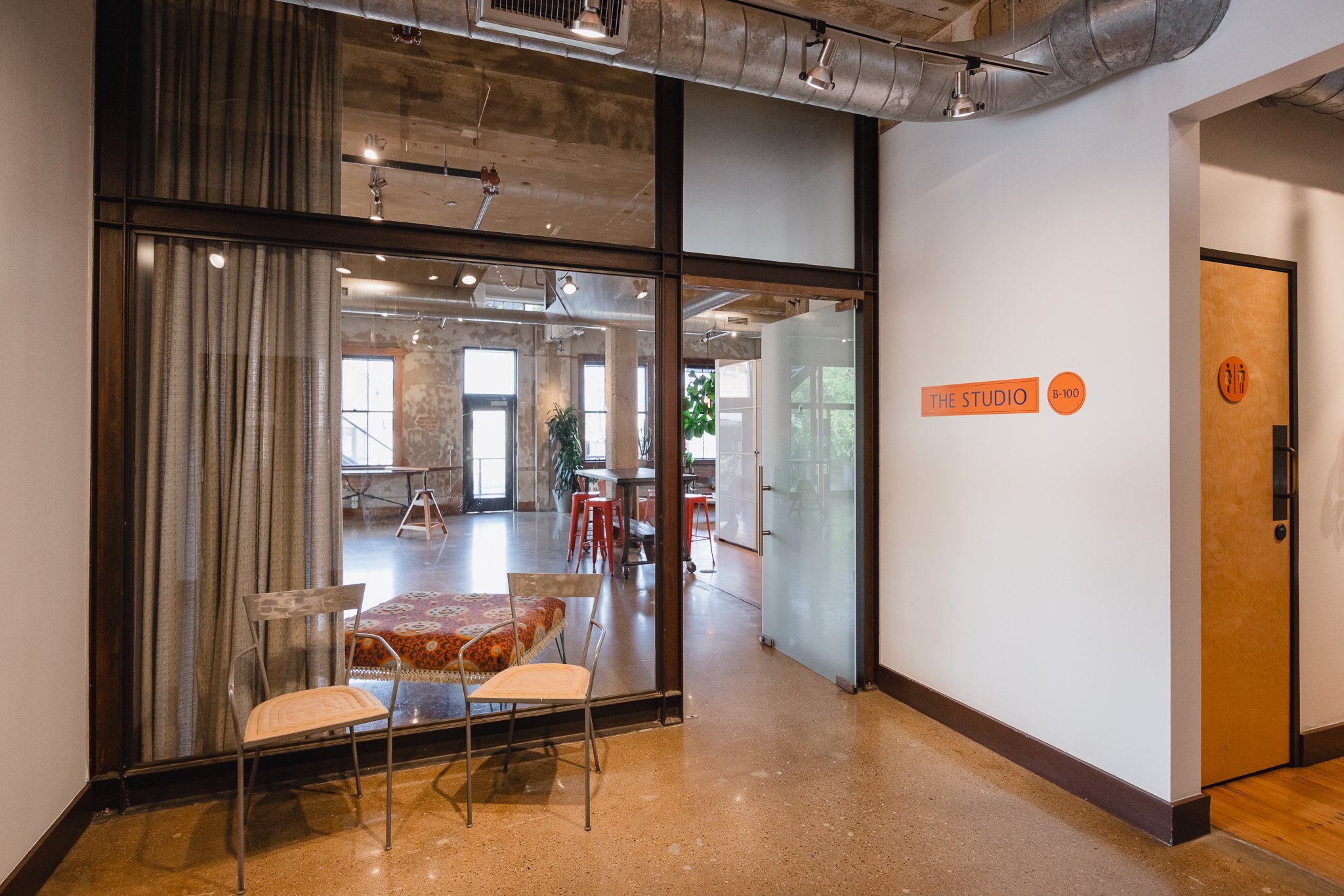Interior view of a modern studio space seen through a large glass window and door, with a small sitting area in the foreground featuring two chairs and a table, and an open room with high windows, exposed brick walls, plants, and furniture in the bac