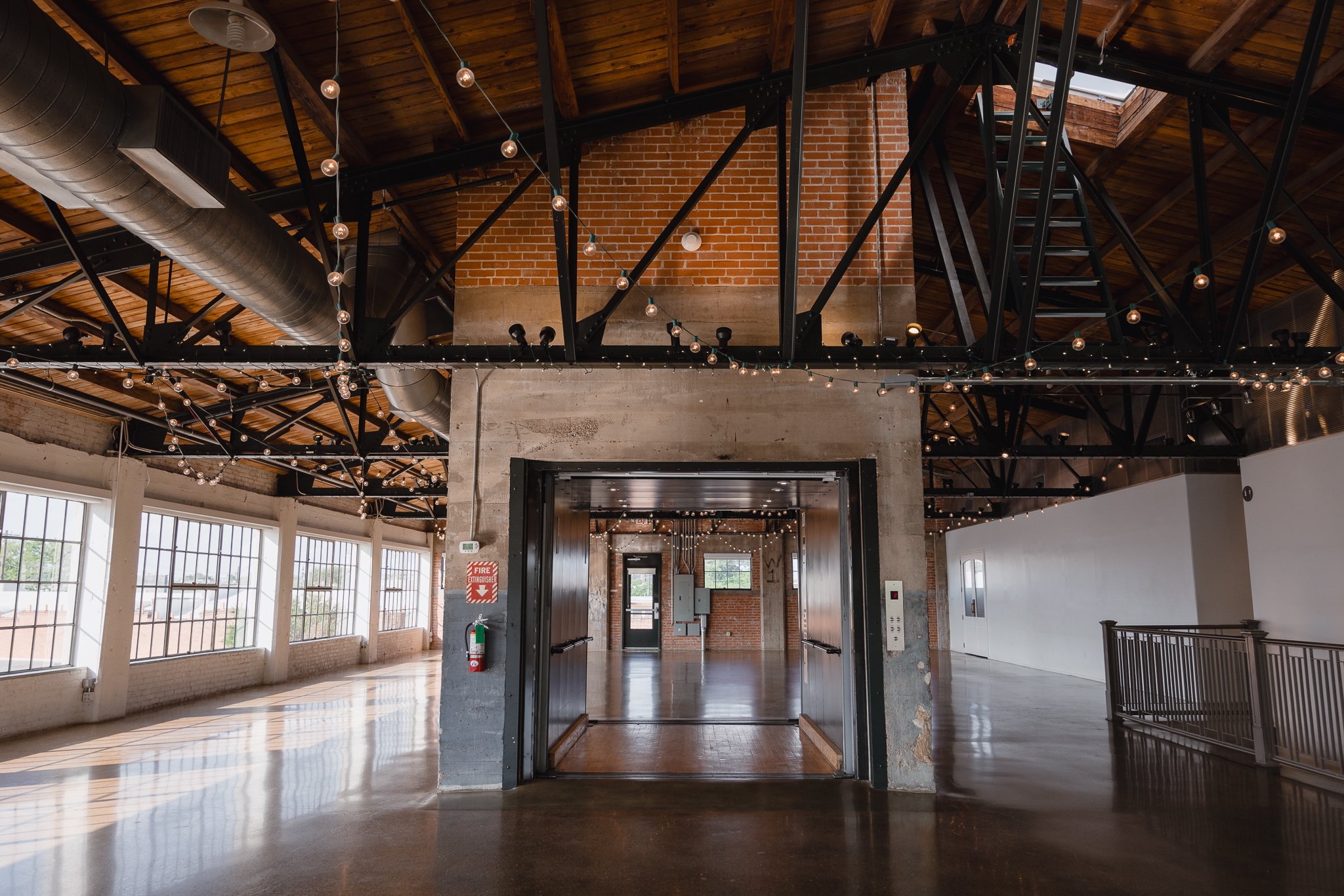 An industrial-style interior space with wooden ceiling, black metal beams, string lights, large windows, a brick and concrete wall, an elevator with open doors, and a polished concrete floor.