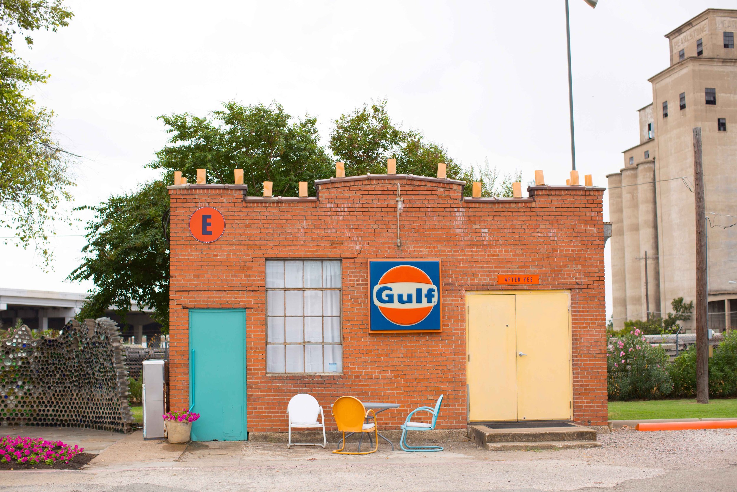 Small brick building with Gulf sign, blue door, yellow door, colorful chairs outside, potted flowers, trees, and an industrial structure in the background.