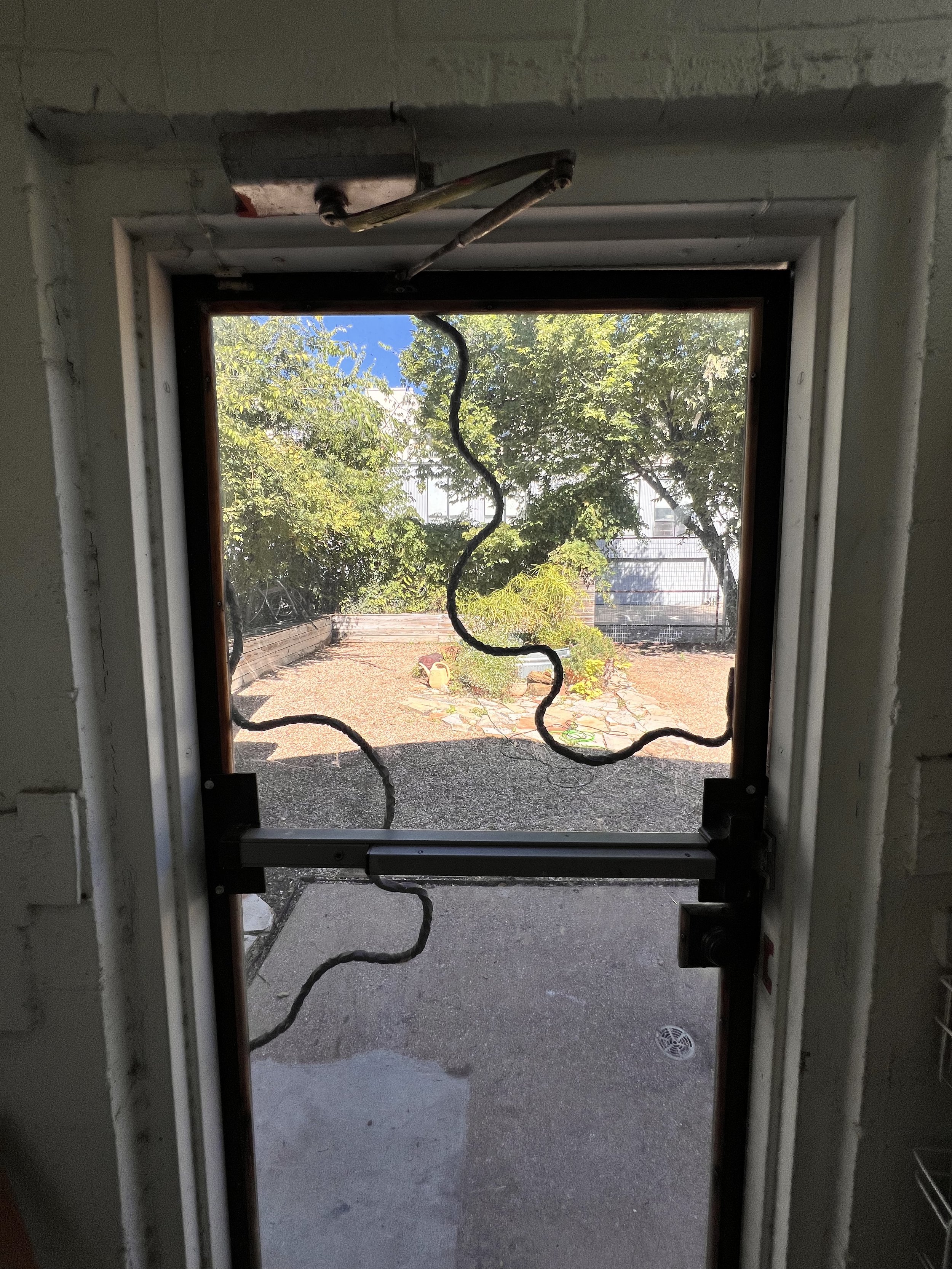 Interior view of a door with broken or removed glass, showing the outdoor yard with trees, plants, and a pathway.