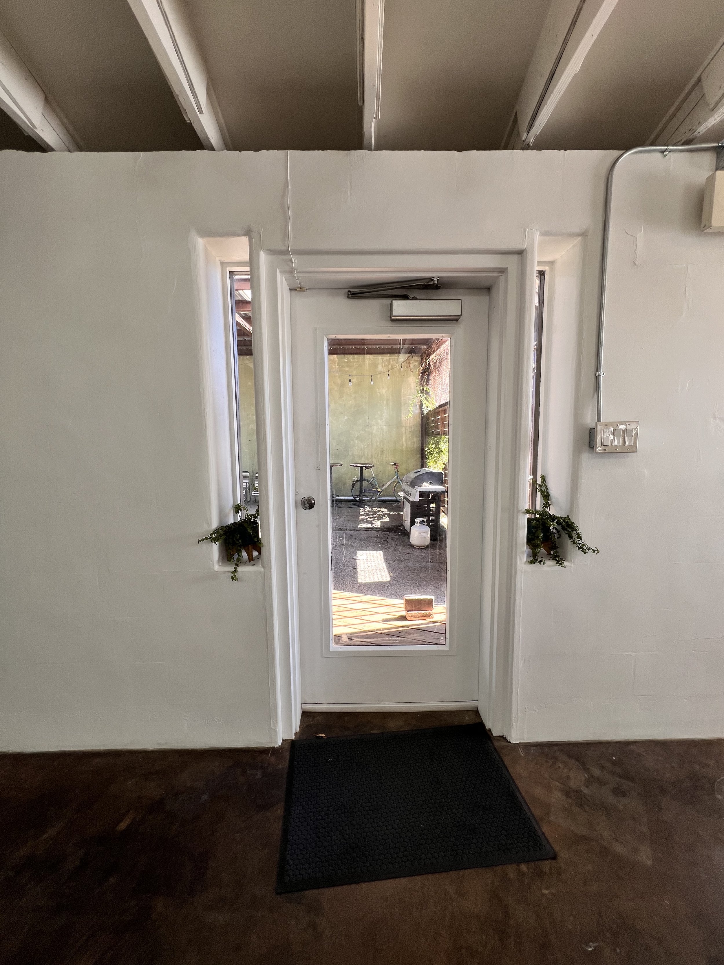 White door with a glass panel leading to an outdoor patio with a BBQ grill and bicycles, flanked by two small windows with planters.