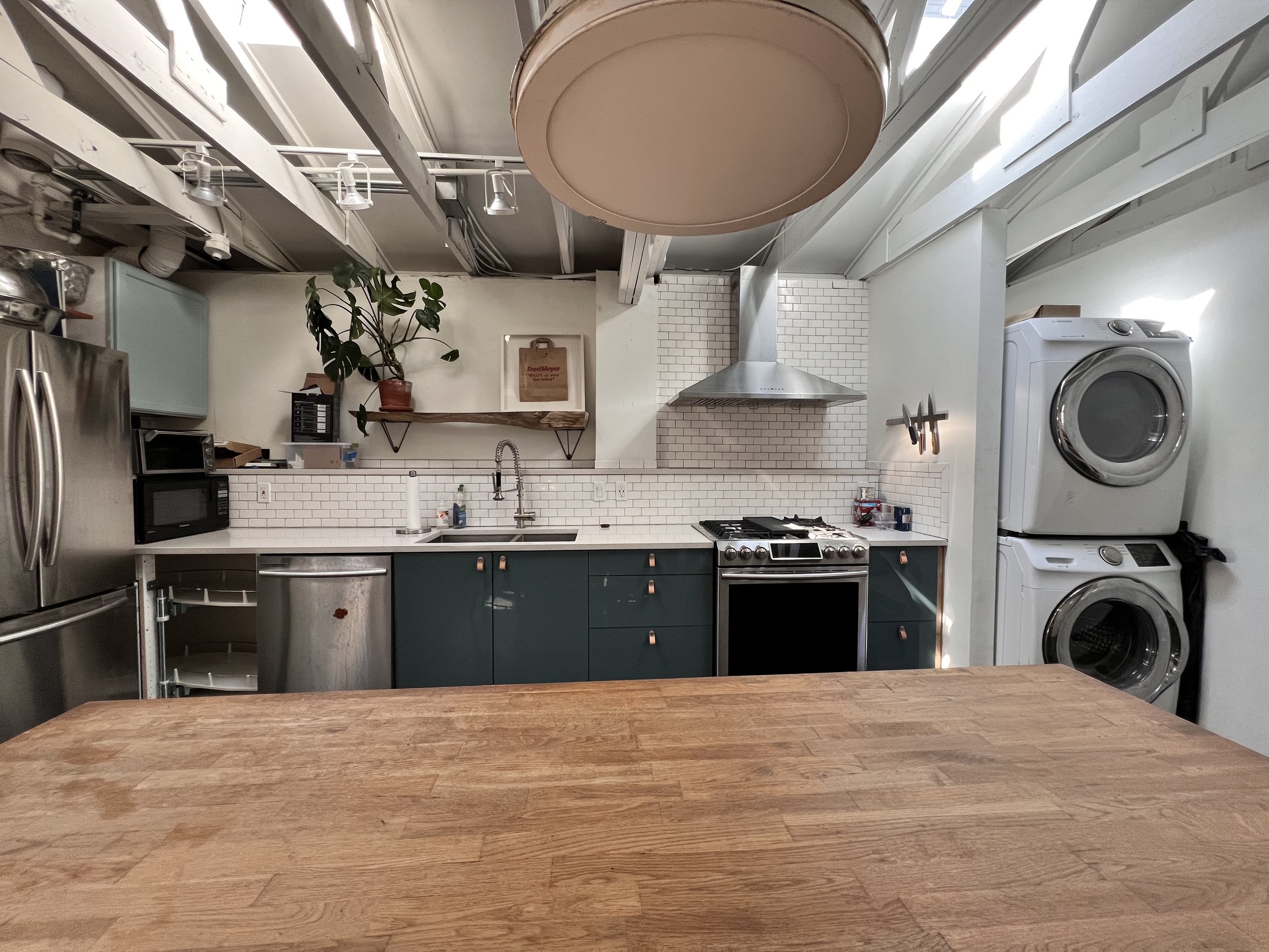 Modern kitchen with wooden countertop in foreground, teal cabinets, white subway tile backsplash, stainless steel appliances including refrigerator, microwave, dishwasher, and oven, with stacked washer and dryer unit on the right. A potted plant on a