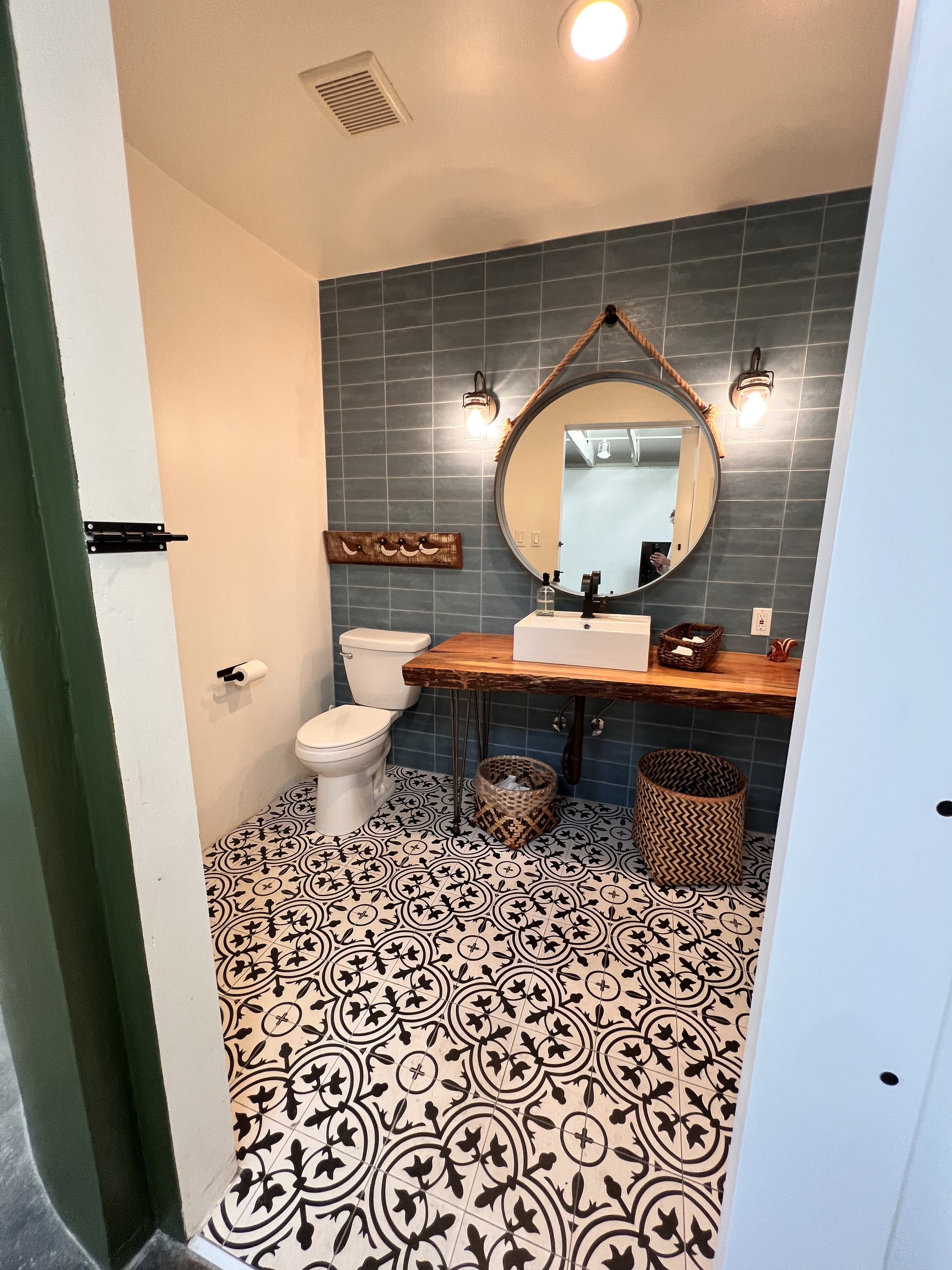 A bathroom with black and white patterned floor tiles, a white toilet, a wooden vanity with a square vessel sink, round mirror with a rope border, and decorative baskets on the floor. The wall behind the sink has greenish-blue tiles, and there are tw