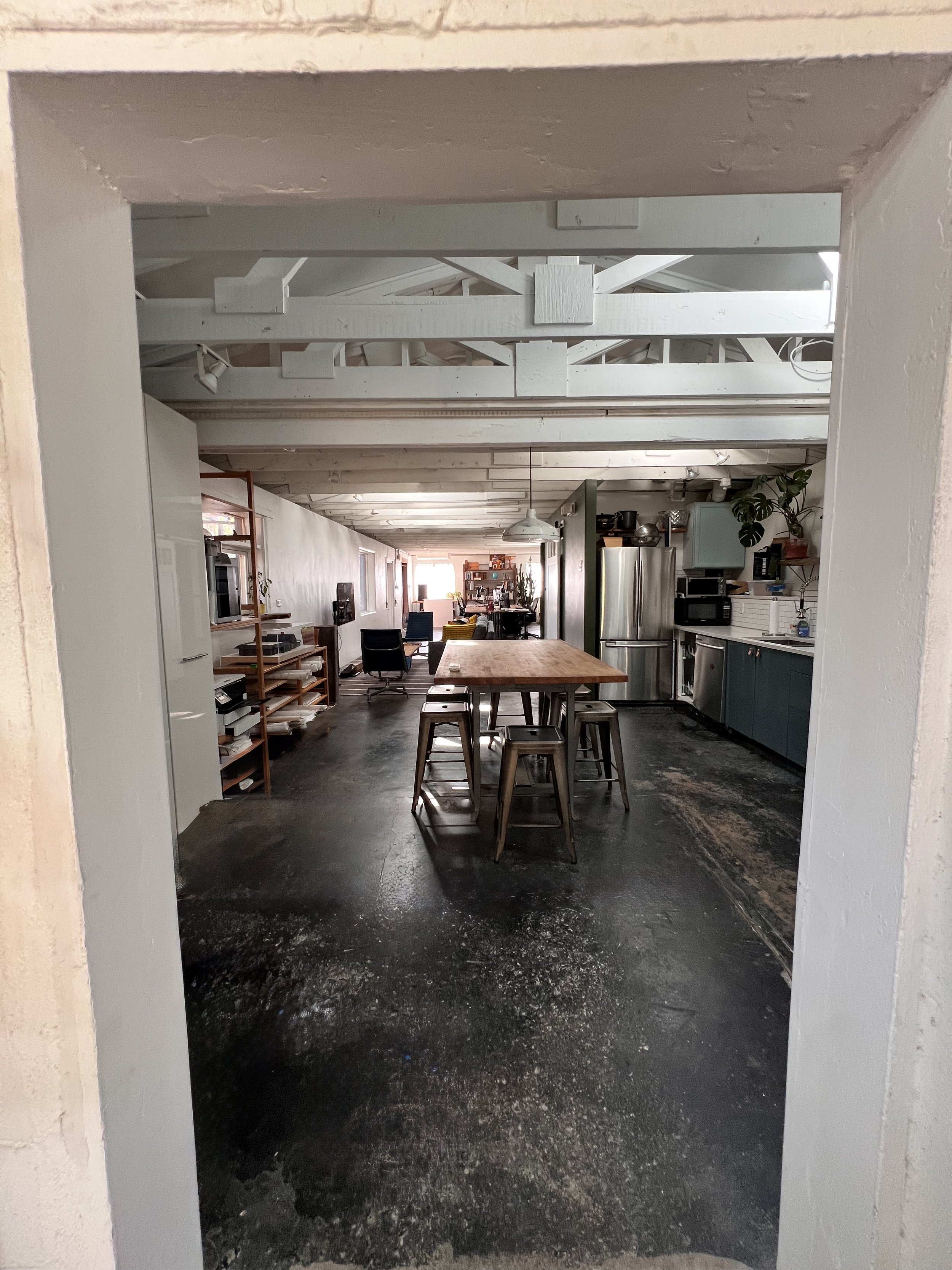 View of a modern open-concept kitchen and living space, with a central wooden dining table, stools, kitchen appliances, and seating area with chairs and a coffee table in the background.