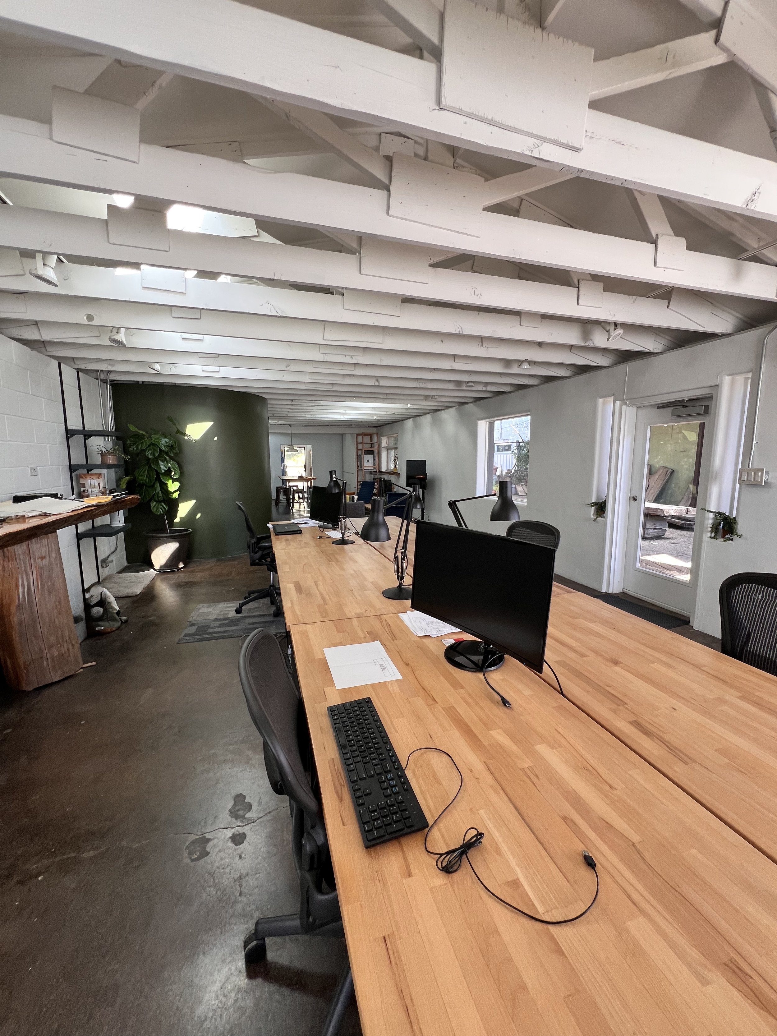 Modern office space with wooden tables, black office chairs, computer monitors, keyboards, and desk lamps. The room has white walls, large windows, and a ceiling with exposed wooden beams painted white.