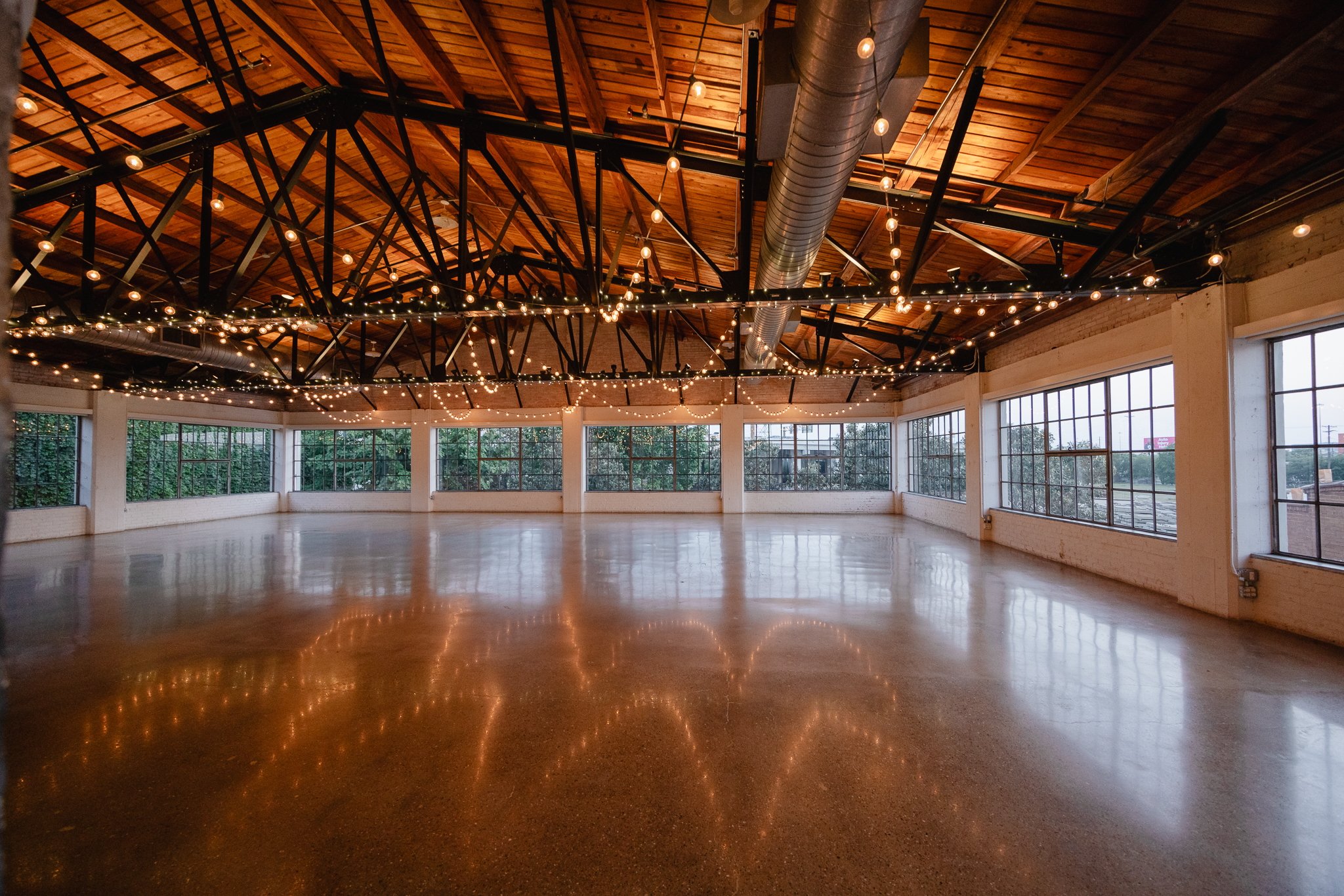 Empty event space with string lights hanging from high, wooden vaulted ceiling, large windows along the walls, and a polished floor reflecting the lights.