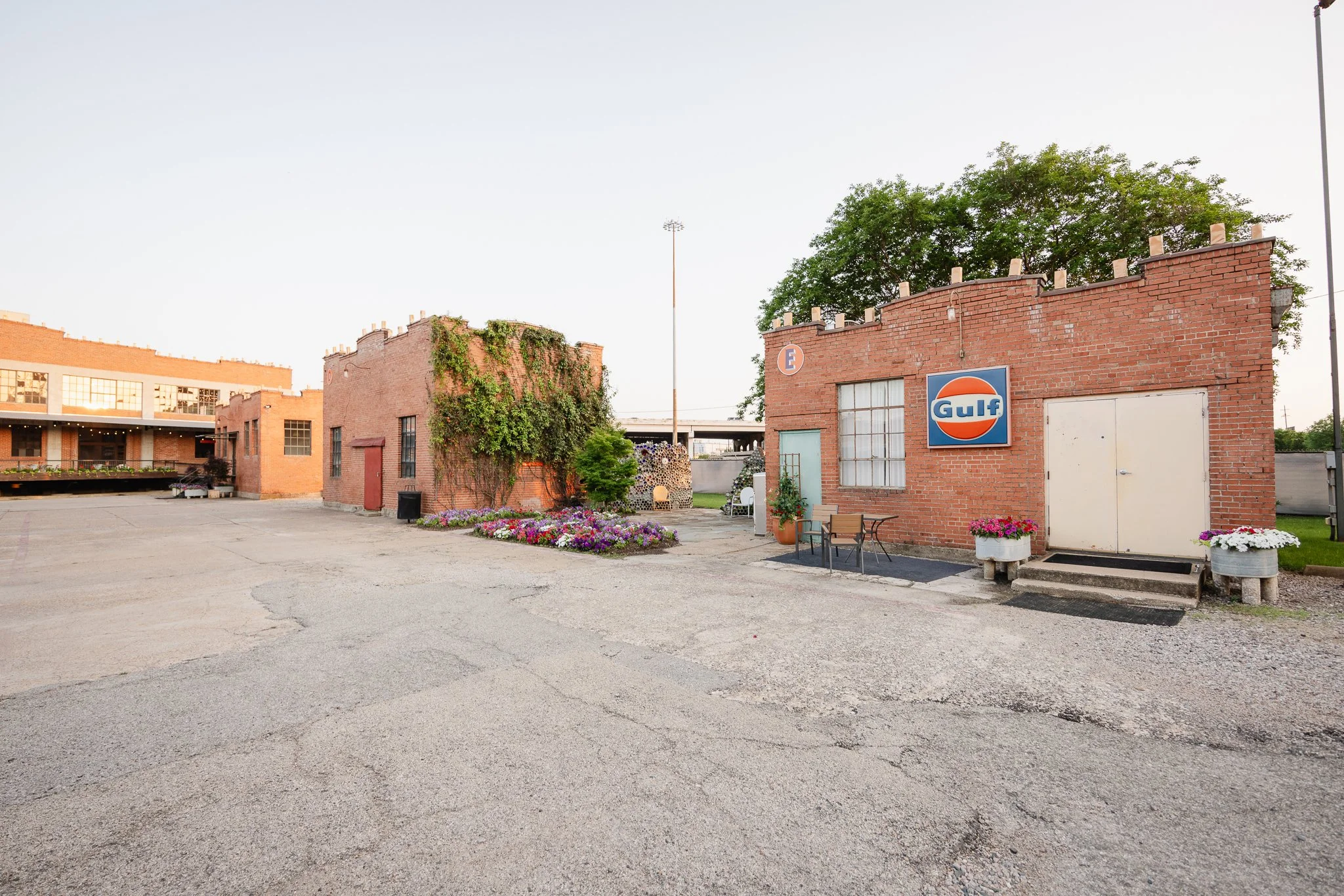 A small brick building with a Gulf oil sign, surrounded by flower pots and outdoor furniture in an open parking lot with cracked pavement, against a clear sky.