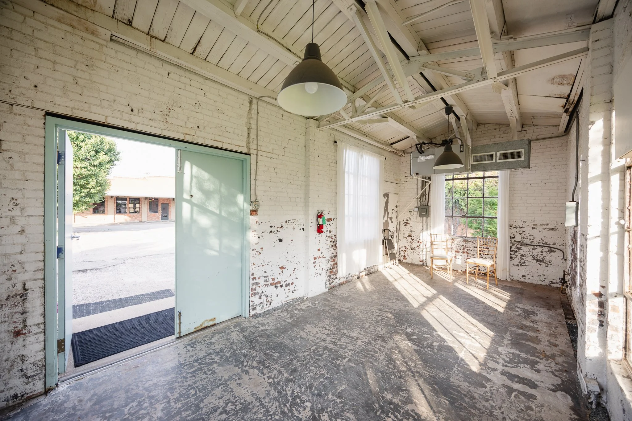 An empty industrial-style room with exposed white brick walls, a concrete floor, and a high ceiling with visible beams. Sunlight streams in through large windows, casting shadows on the floor. There are two chairs near the window and hanging industri