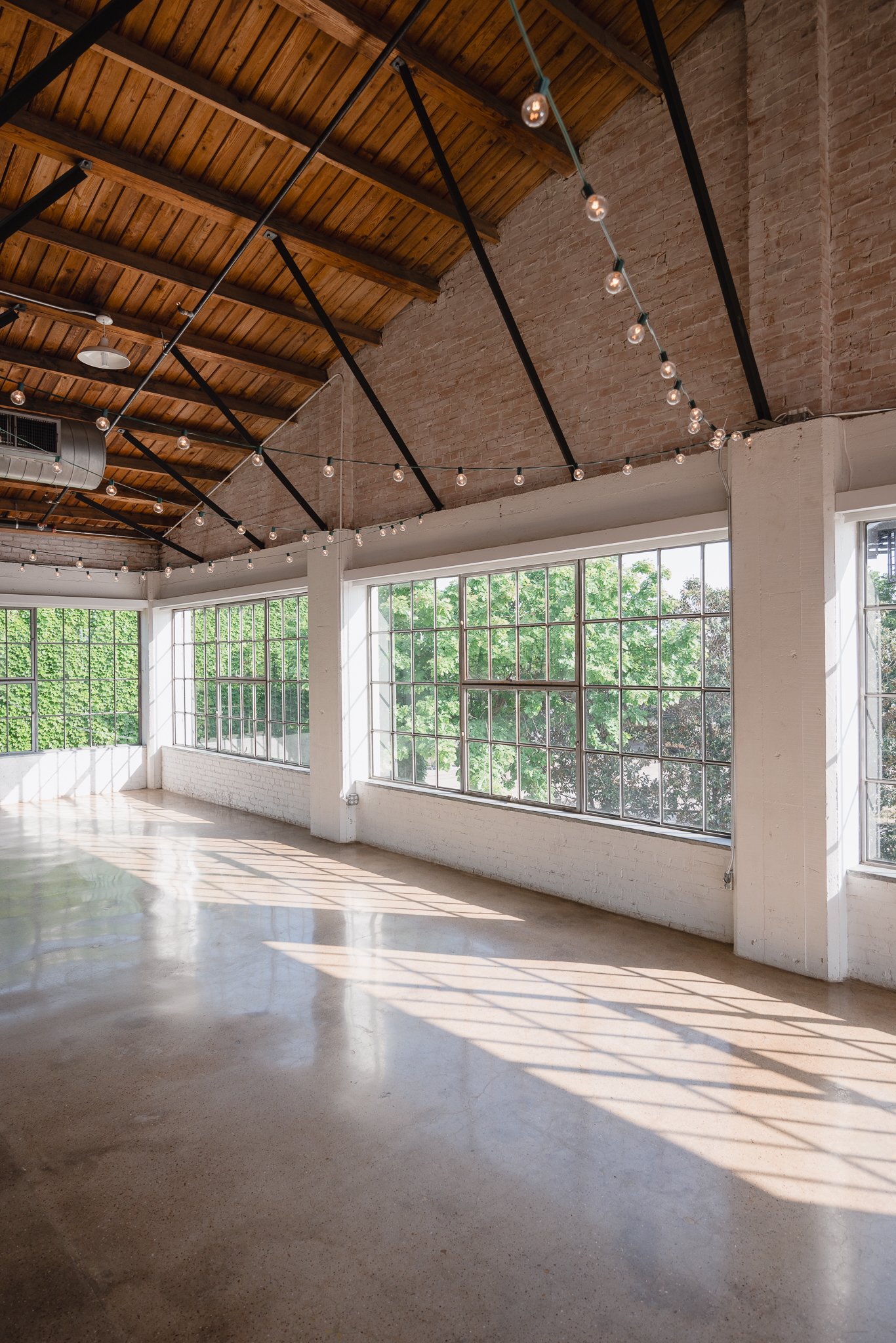Bright, spacious indoor area with large windows, exposed brick walls, polished concrete floor, and string lights hanging from a wooden ceiling.