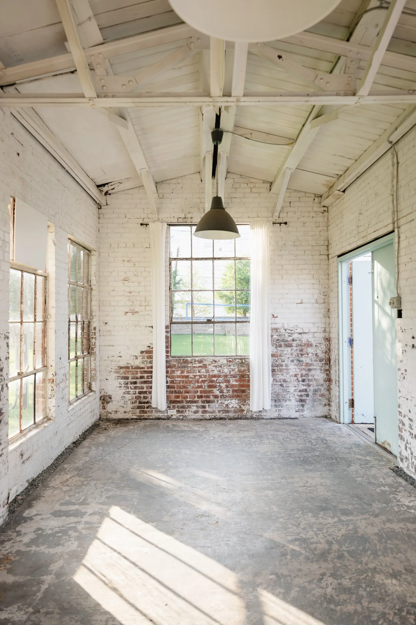 An empty room with white brick walls, large windows, and a high sloped ceiling with exposed beams, painted white. There is a single black hanging pendant lamp in the center, and sunlight streams through the windows, casting shadows on the floor.