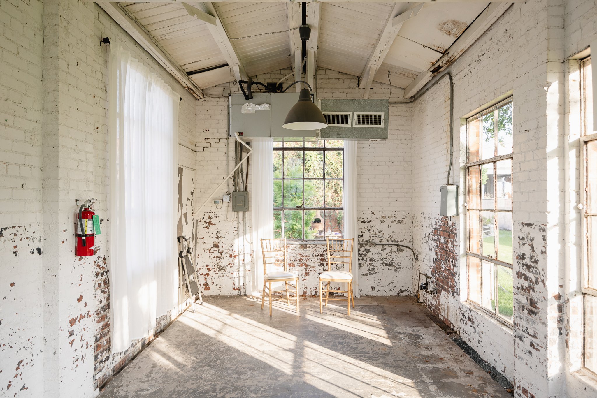 Empty room with white brick walls, a large window with sheer curtains, two chairs, a fire extinguisher on the wall, and an industrial-style hanging light fixture.