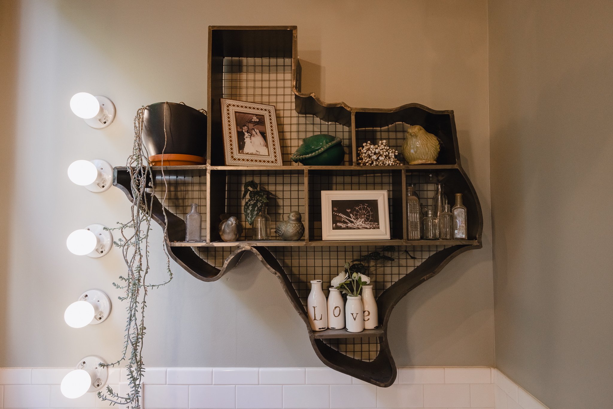 Decorative wall shelf with various items including photo frames, glass bottles, vases, a bag, and small ornamental objects, next to a set of four white light bulbs mounted on the wall