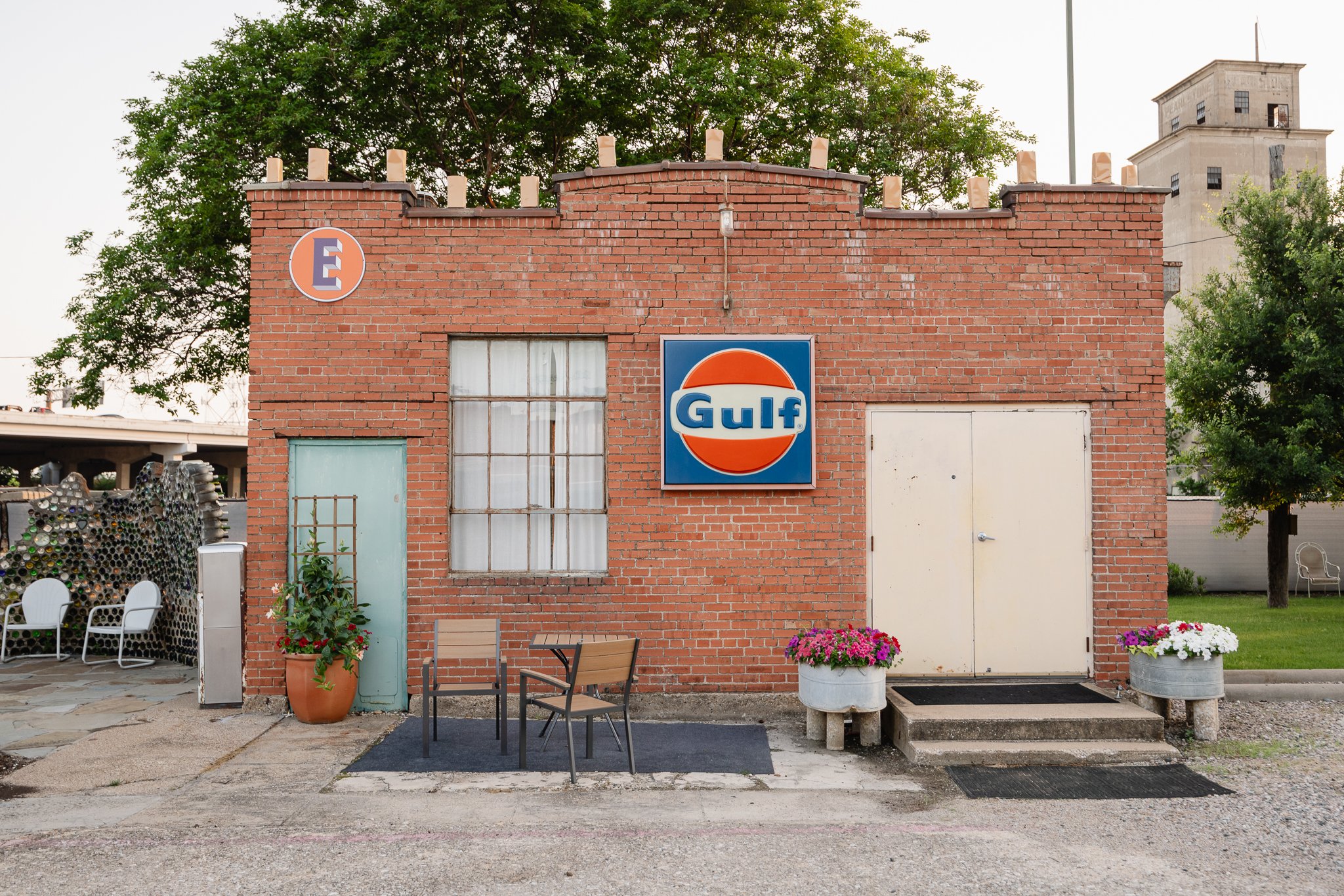Small red brick building with Gulf oil sign, two doors, and a window, surrounded by potted flowers and outdoor seating, likely a vintage gas station or retro storefront.