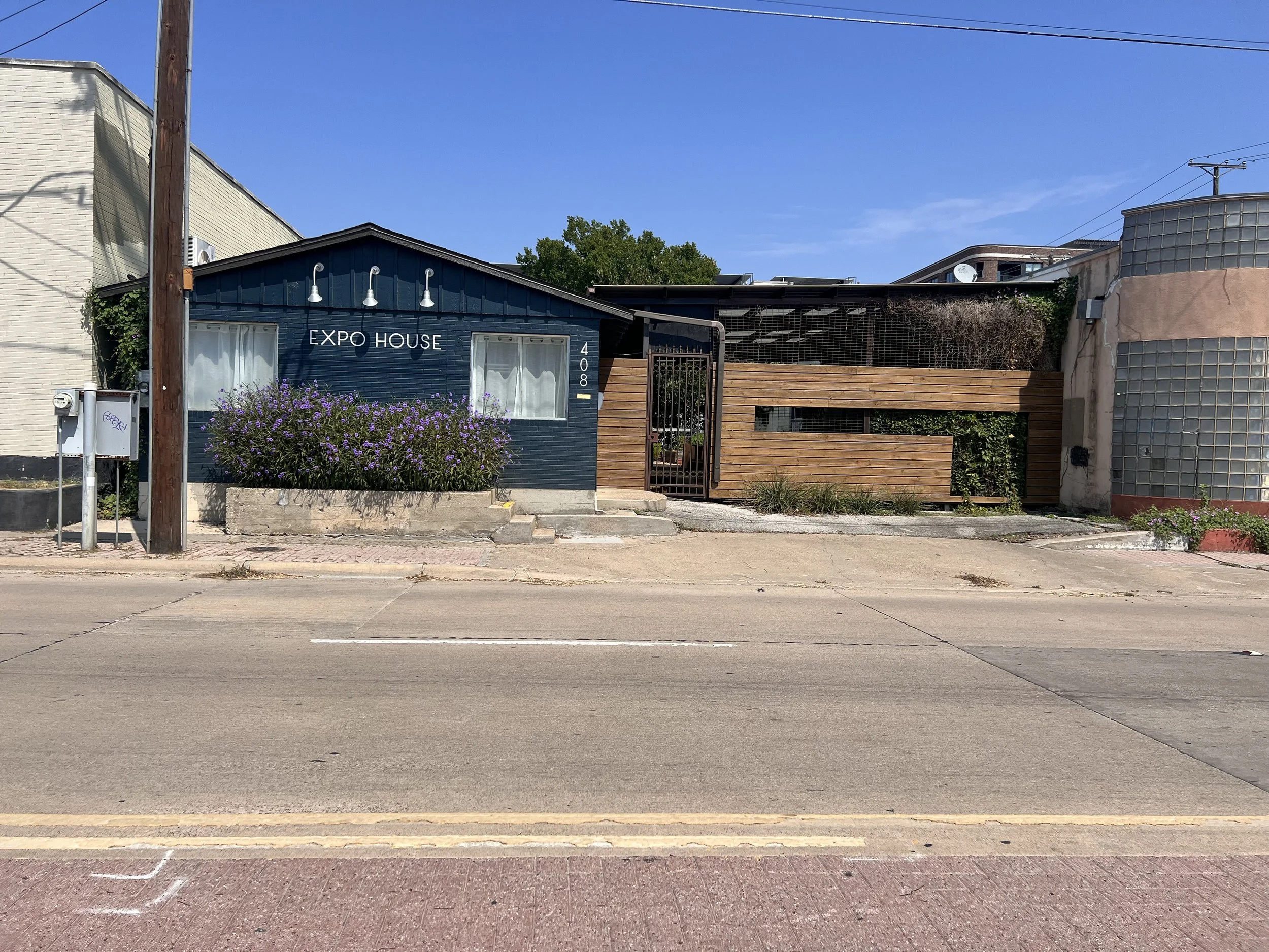 A small building painted blue with a sign that says 'EXPO HOUSE' and the street number 408, surrounded by a concrete sidewalk and street, with purple flowers in front and a wooden and glass fence to the right.