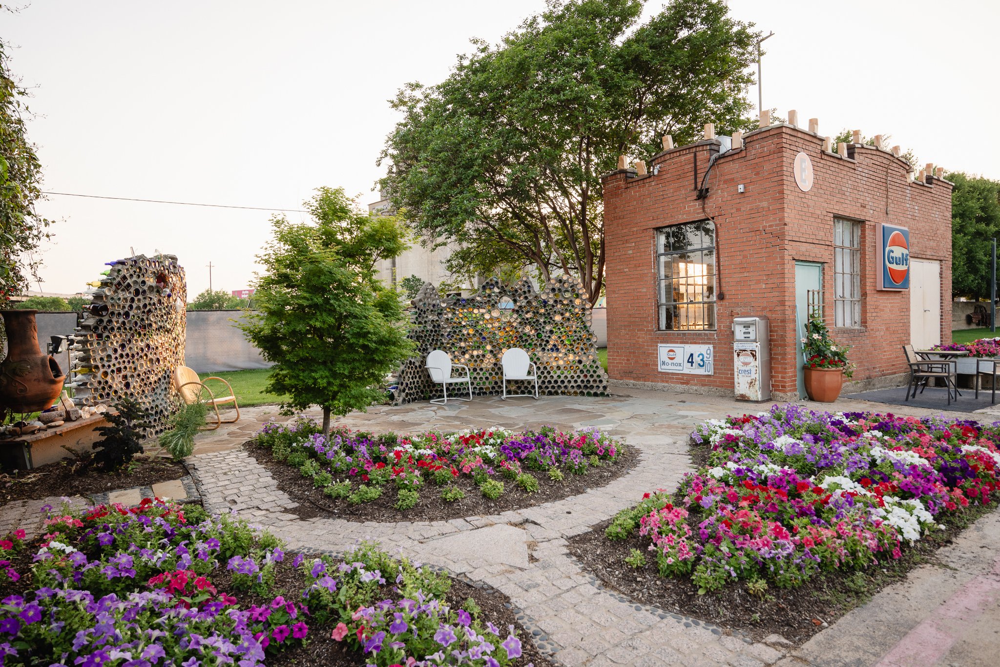 A small brick building with a Gulf gas station sign, surrounded by colorful flower beds and garden decorations, including chairs and art made from recycled materials.
