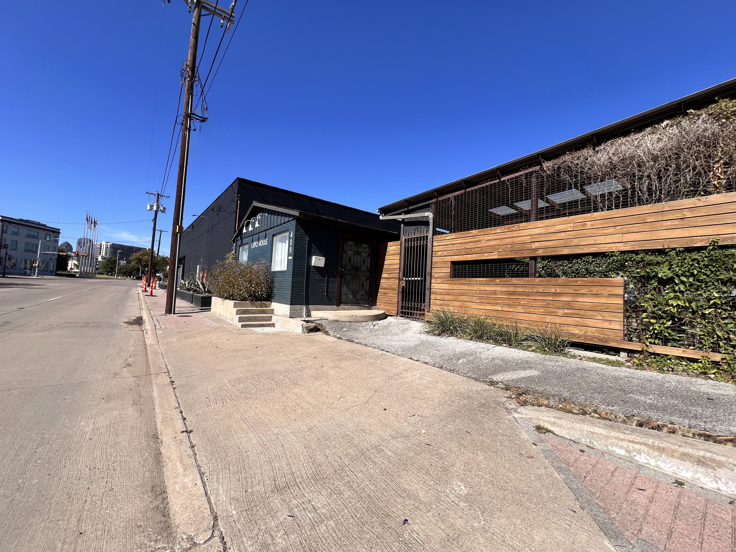 A quiet city sidewalk with utility poles and buildings, including a small dark-colored building with the sign 'Exo House' and a modern wooden fence, under a clear blue sky.
