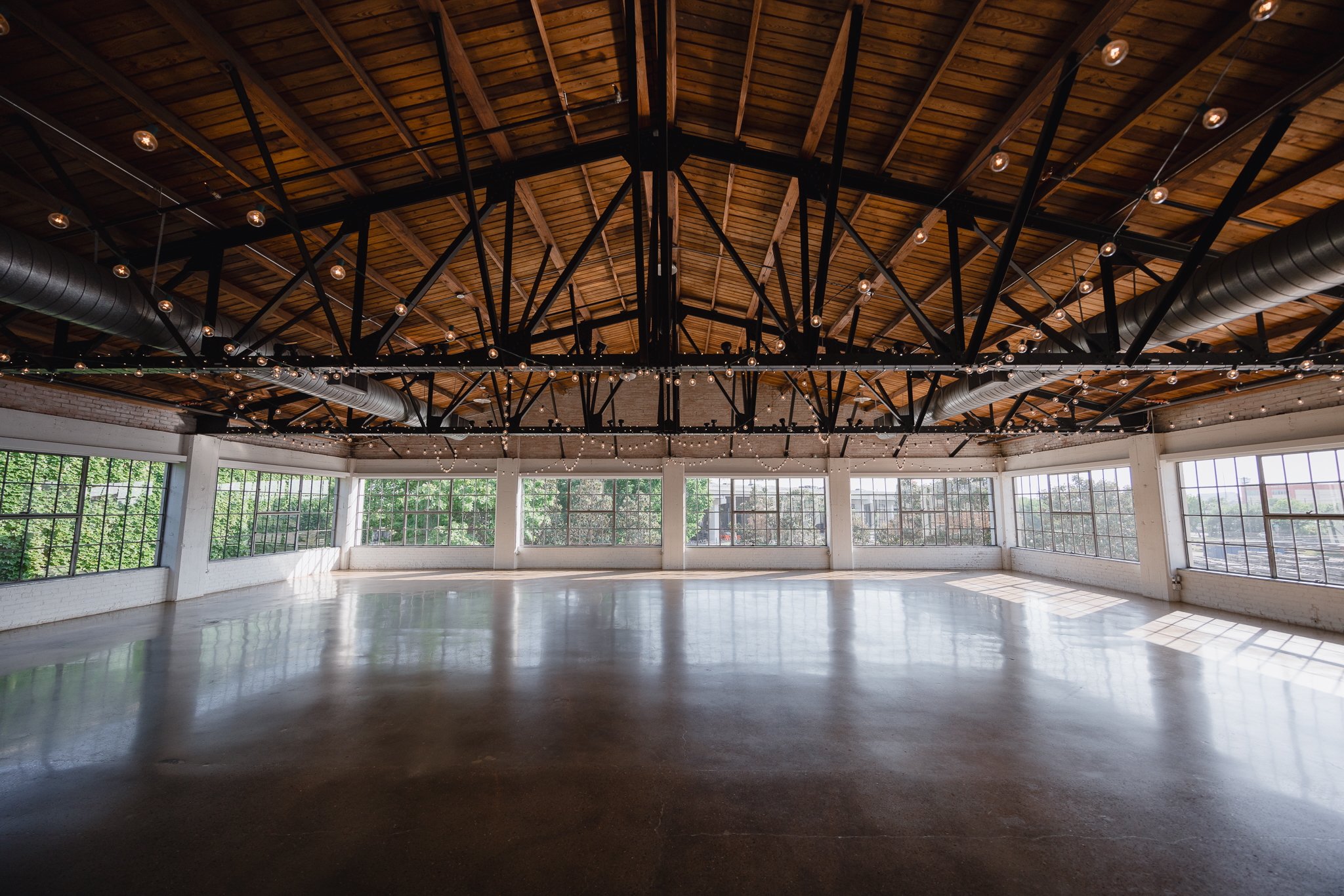 Empty event space with large windows, polished concrete floors, exposed brick walls, and string lights hanging from the high wooden ceiling.