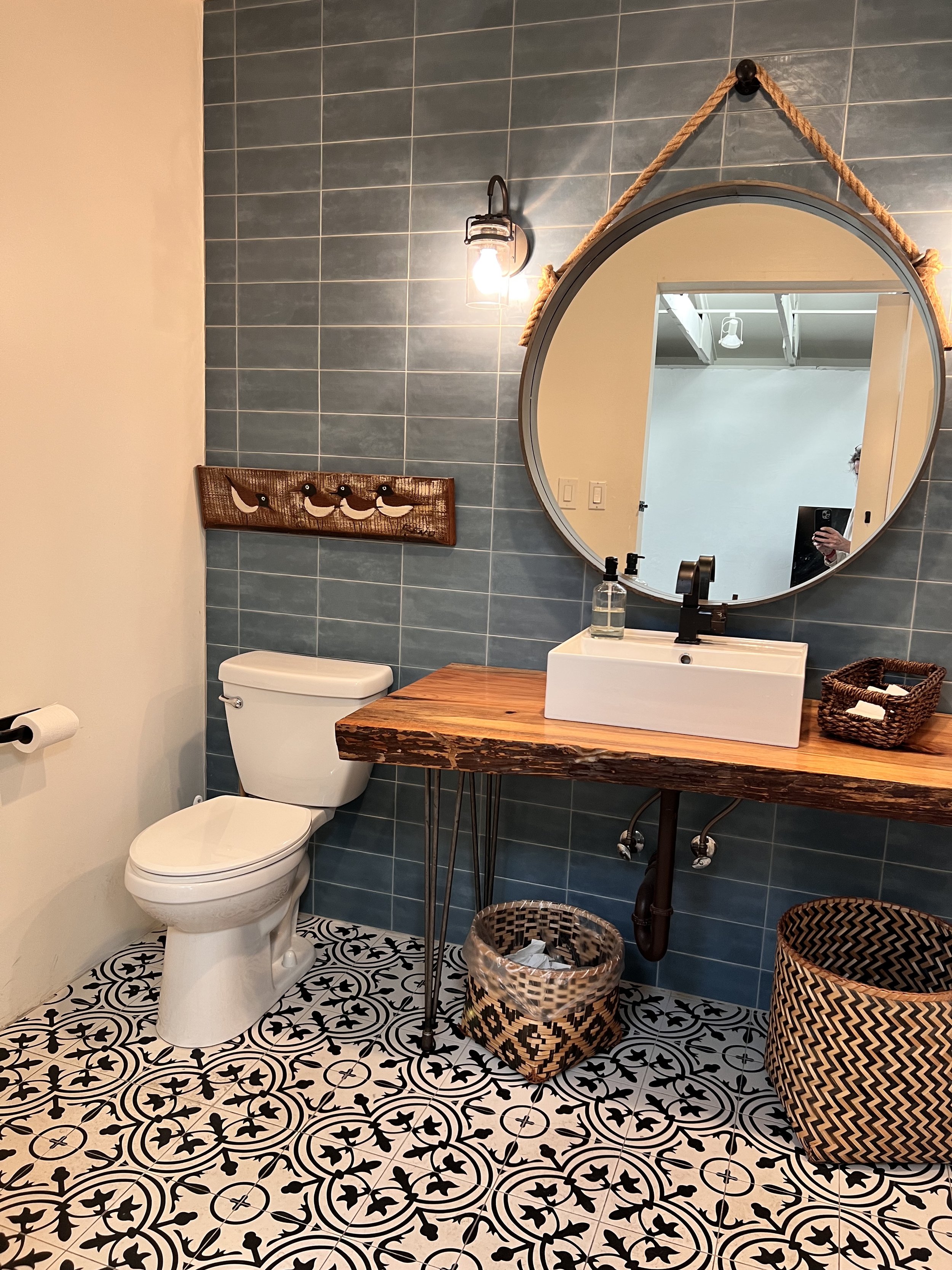 Bathroom with blue tiled wall, white toilet, wooden vanity, circular mirror, wicker baskets, patterned black and white floor tiles, and a wall light fixture.