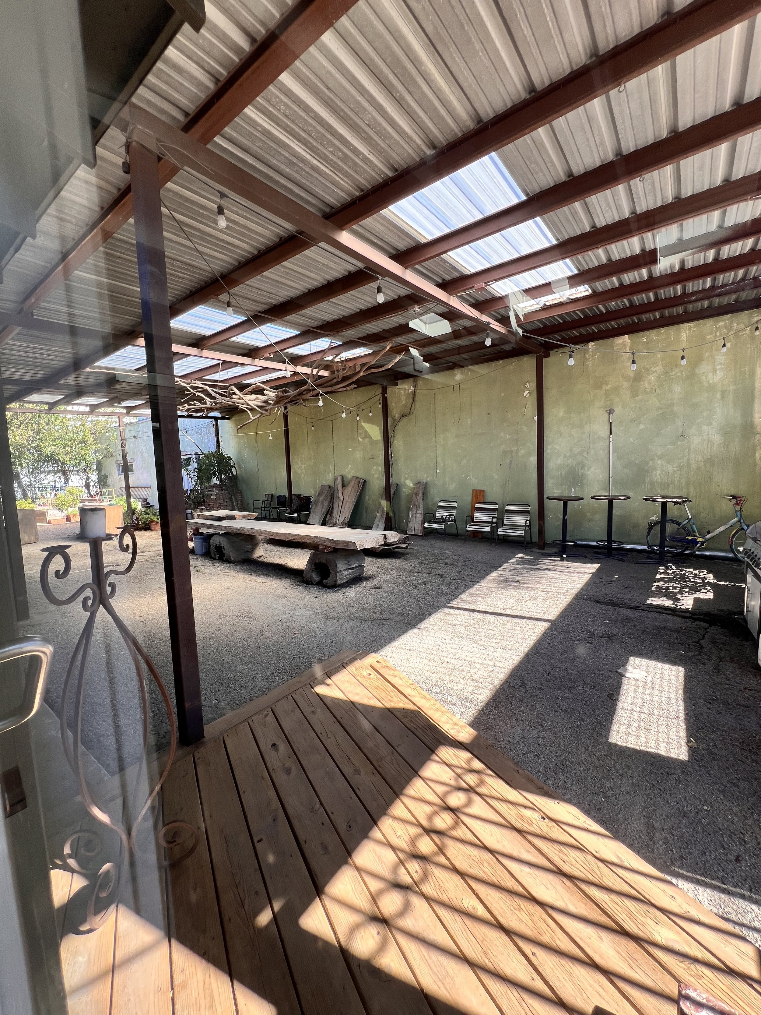 The image shows a partially enclosed outdoor patio area with a metal roof, some chairs against a wall, a wooden table with logs underneath, and some bicycles in the corner. The floor is a mix of wooden decking and gravel, with sunlight casting shadow
