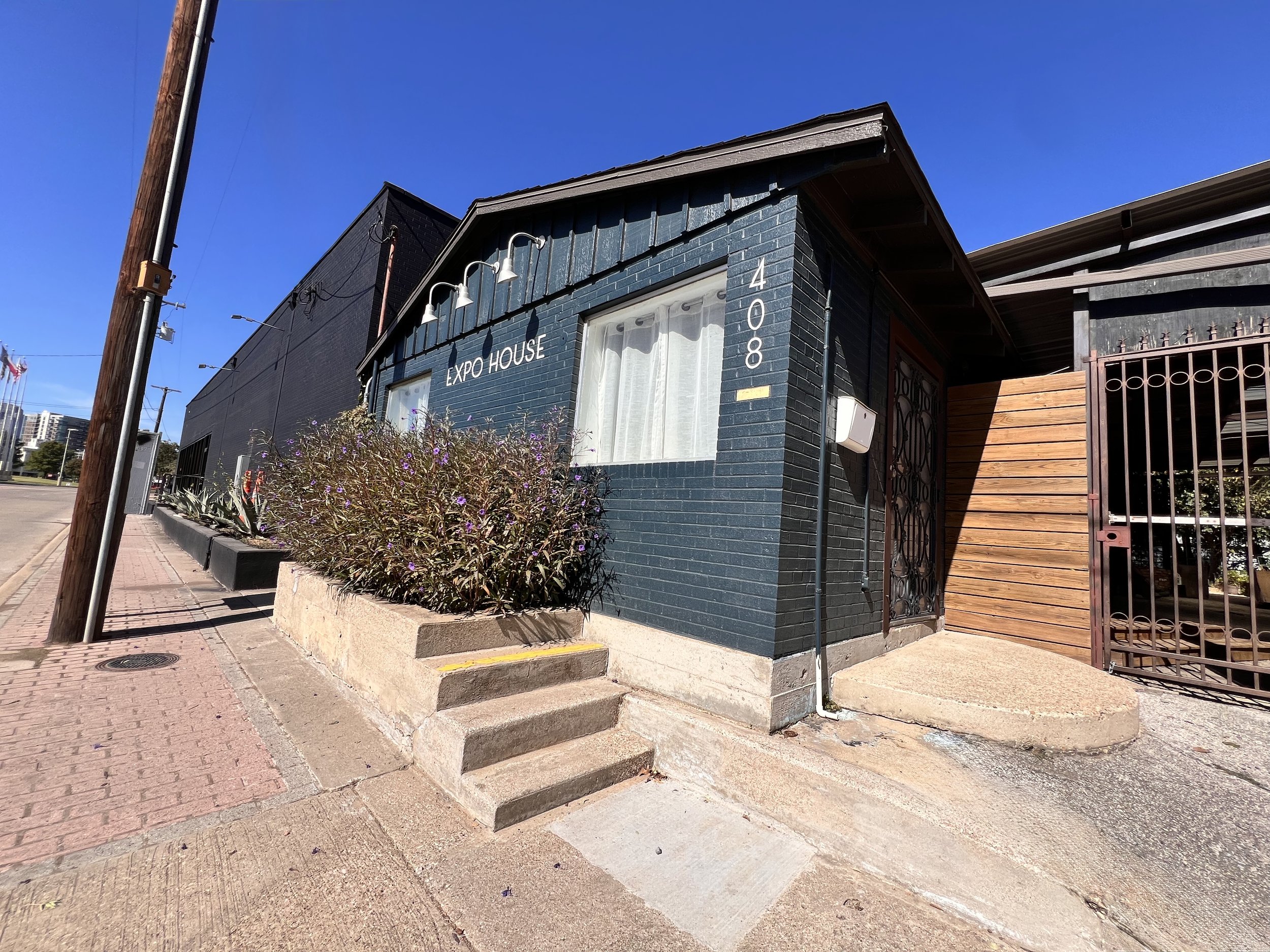 A small black building with white trim labeled 'Expo House' at 408, with gravel steps leading up to a door and a flower bed with purple flowers in front. A streetlamp and power pole are nearby, with a blue sky and city buildings in the background.